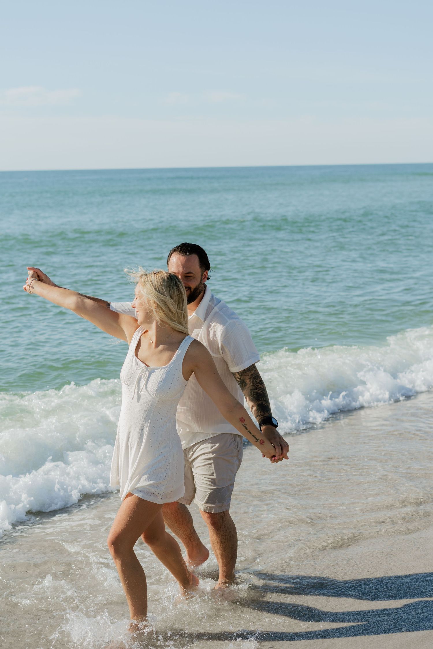 Couple in white outfits holding hands and splashing in ocean waves on a sunny beach.