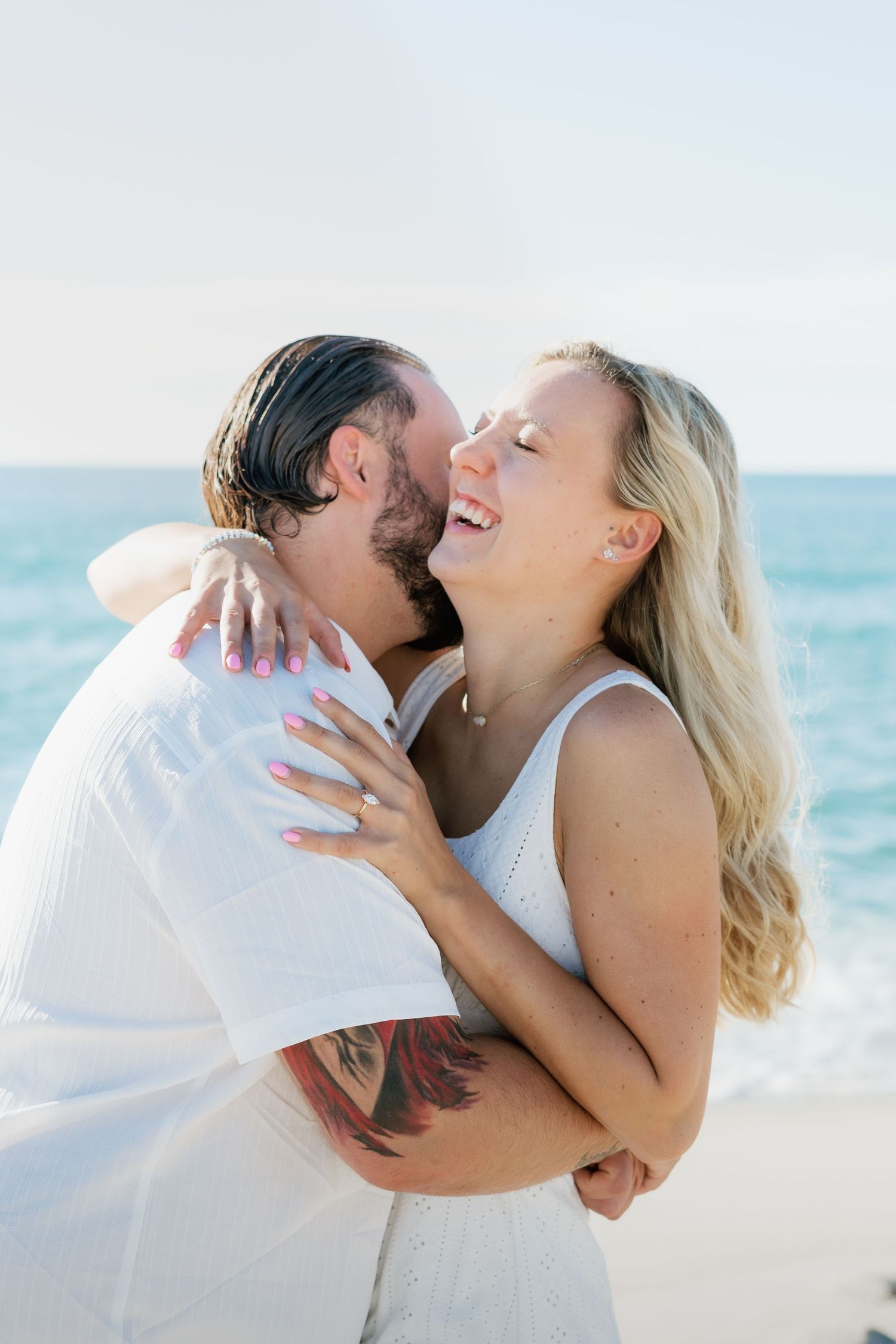 Happy couple embracing and laughing together on a sunny beach with ocean waves in the background.