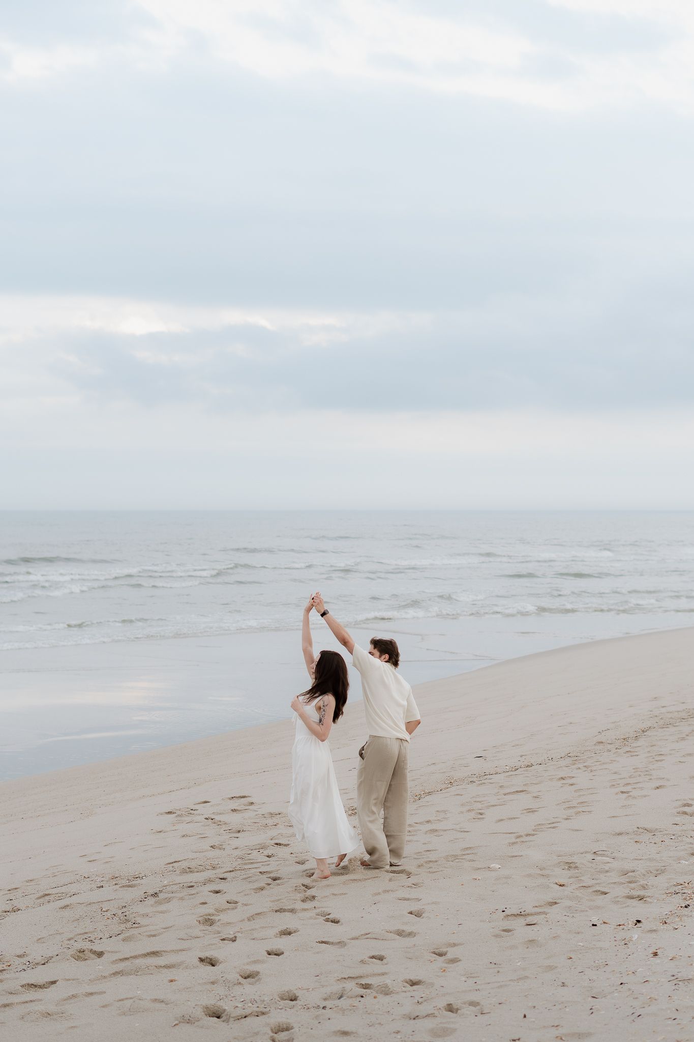 A couple dances joyfully on a sandy beach at dusk, holding hands raised, dressed in white and beige attire.
