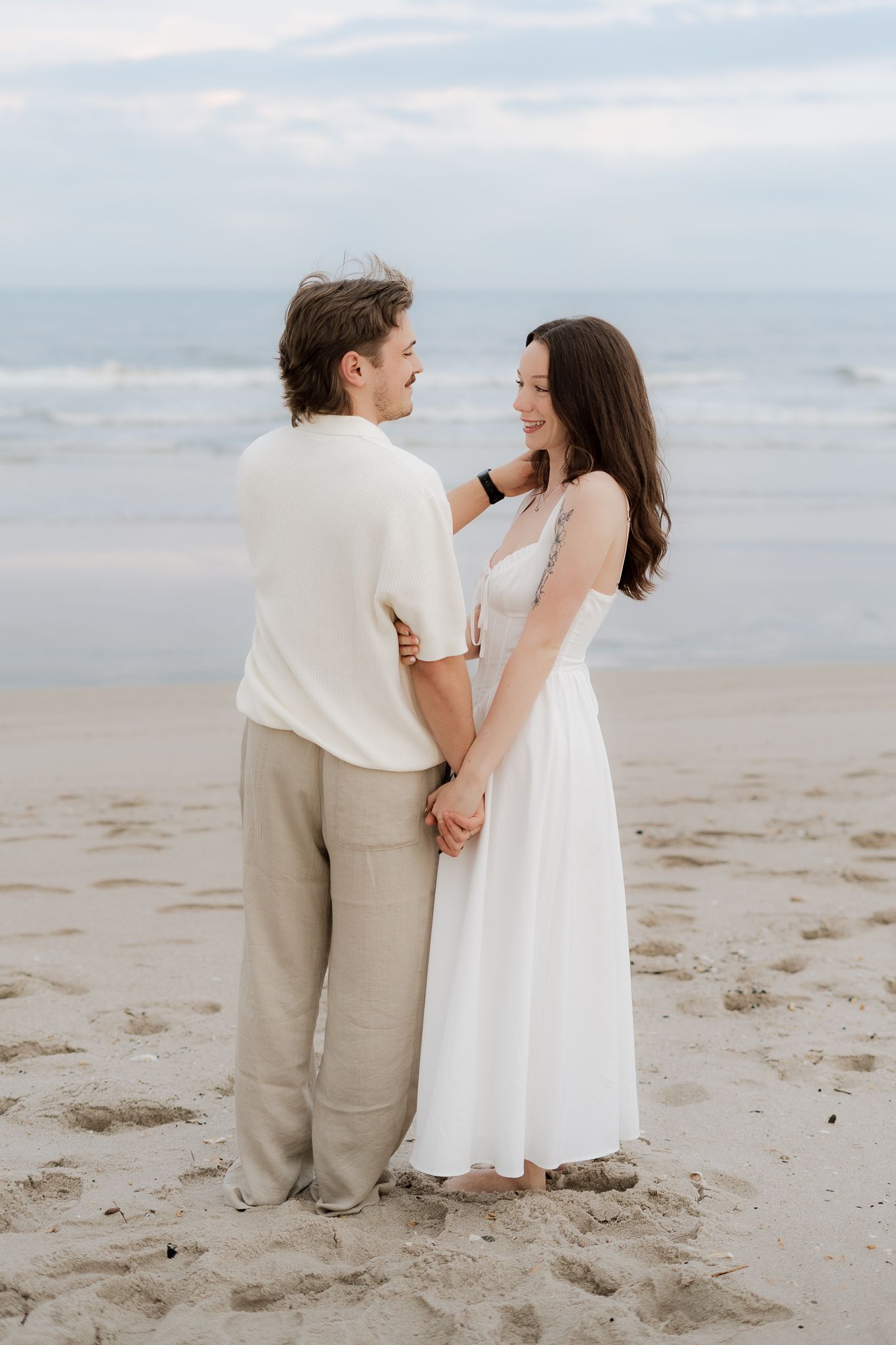 Couple standing on sandy beach holding hands, woman in white maxi dress, man in cream shirt and tan pants, ocean behind.