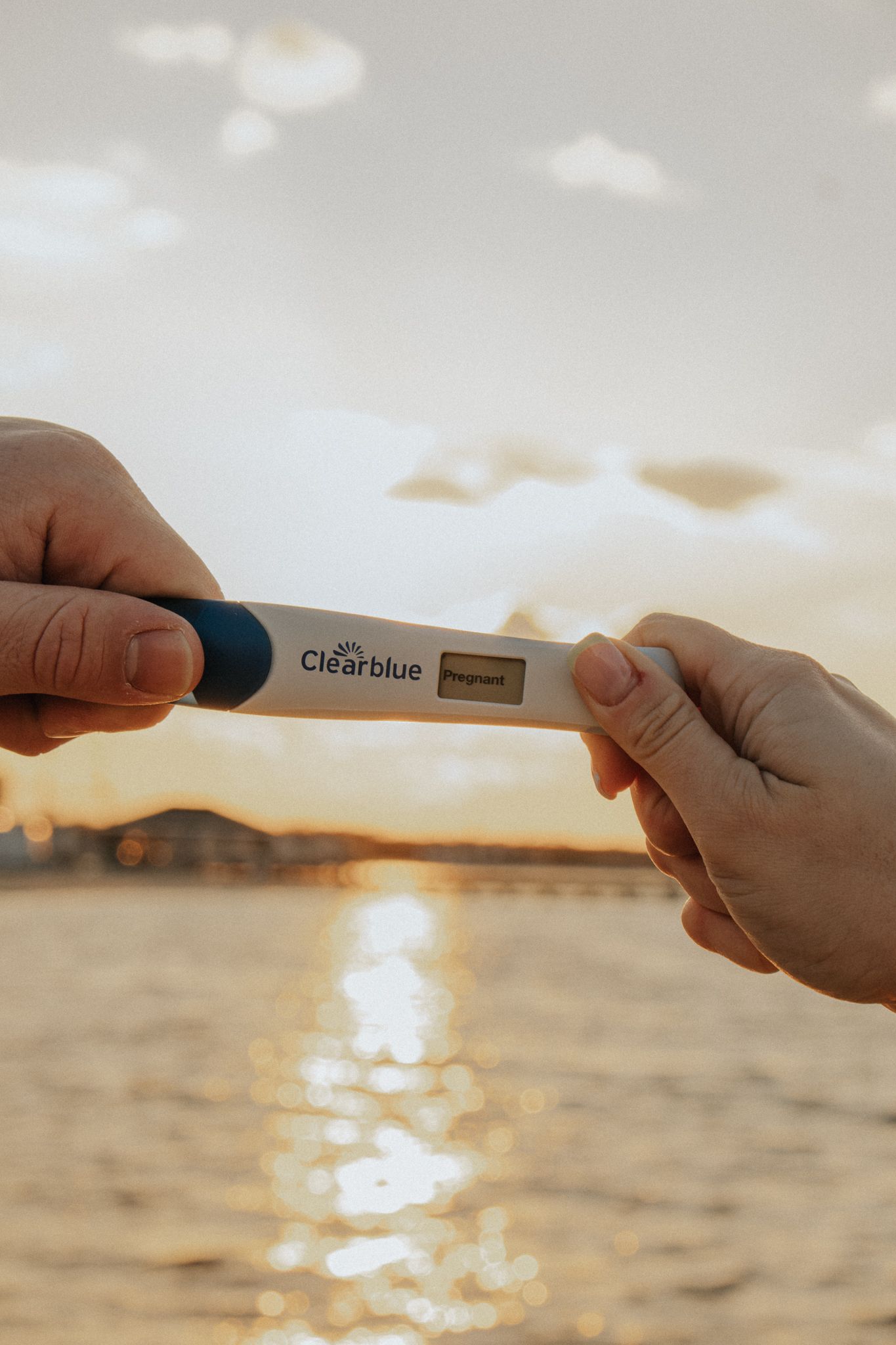 Two hands holding a Clearblue pregnancy test showing pregnant result against a golden sunset over water.