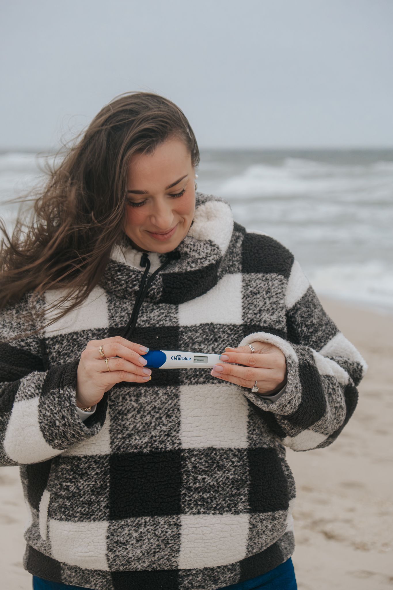 Woman in plaid fleece jacket smiling at pregnancy test on windy beach with ocean waves in background.