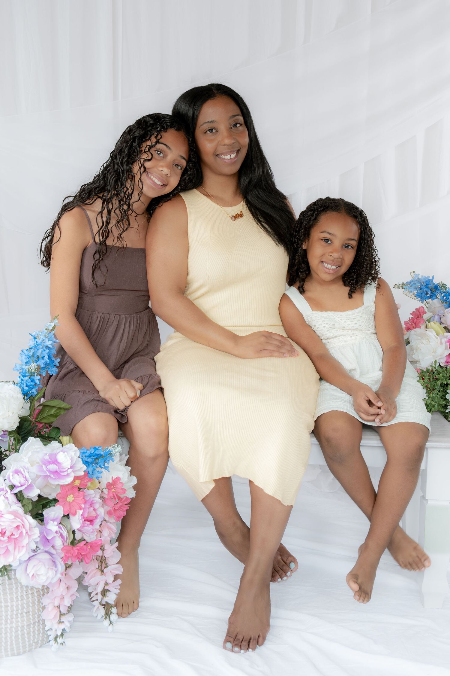 Three women in a white studio with colorful flowers, wearing cream, brown, and white dresses, smiling warmly together.