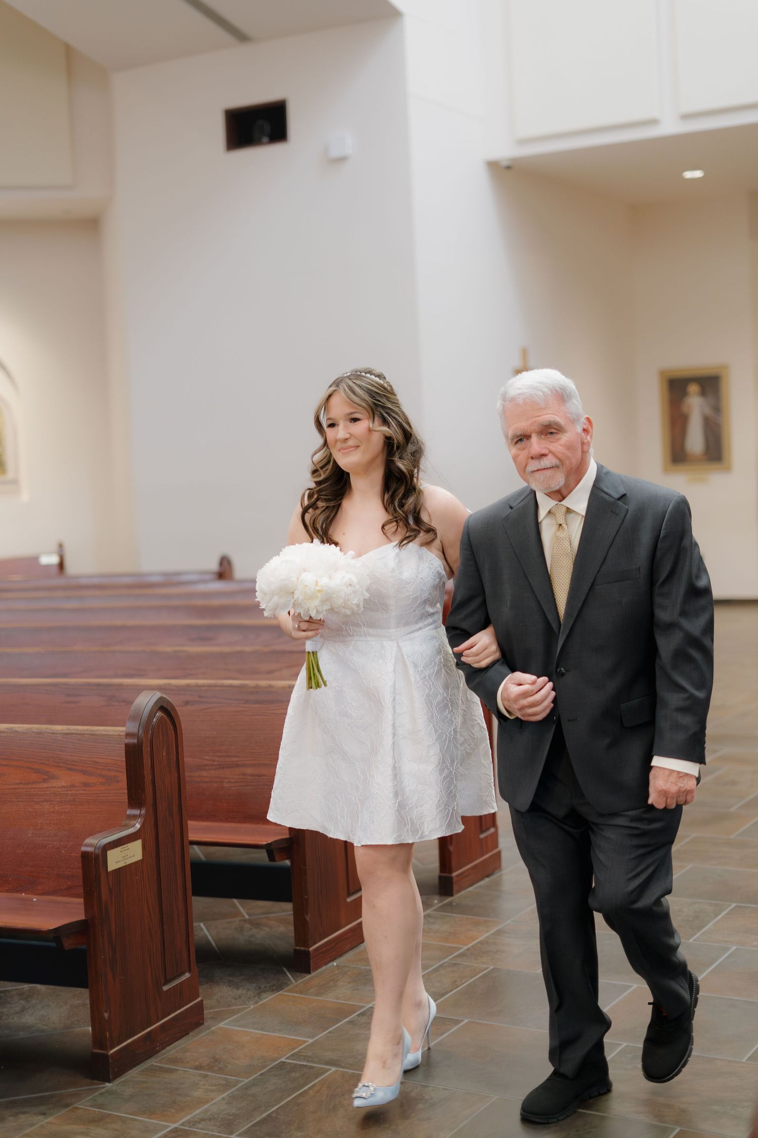 Bride in short white dress walks down church aisle with older gentleman, carrying white floral bouquet.