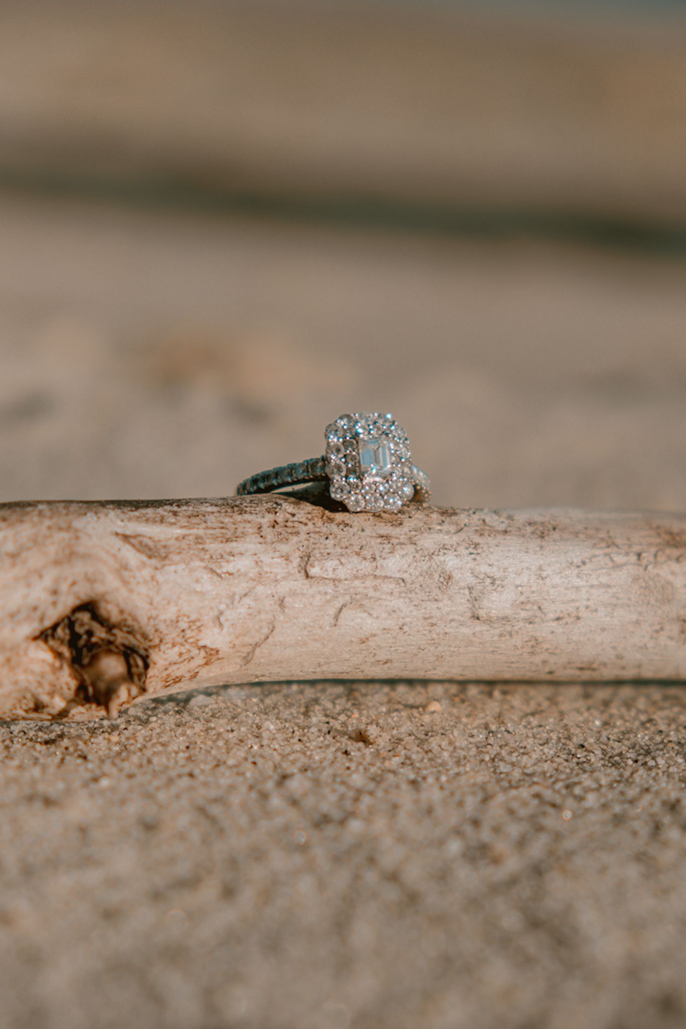 Diamond halo engagement ring with emerald cut center stone displayed on driftwood in sand.