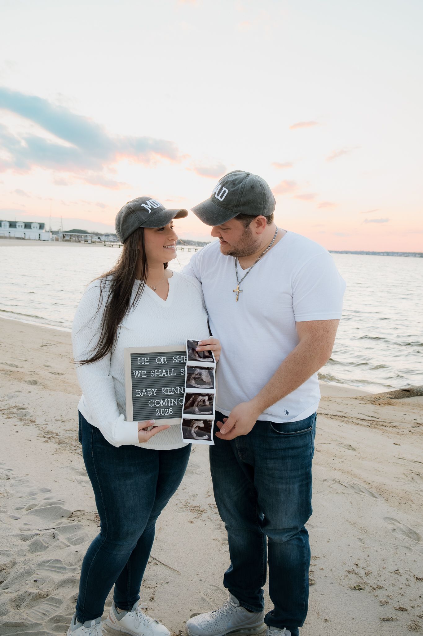 Couple holding pregnancy announcement sign on beach at sunset.
