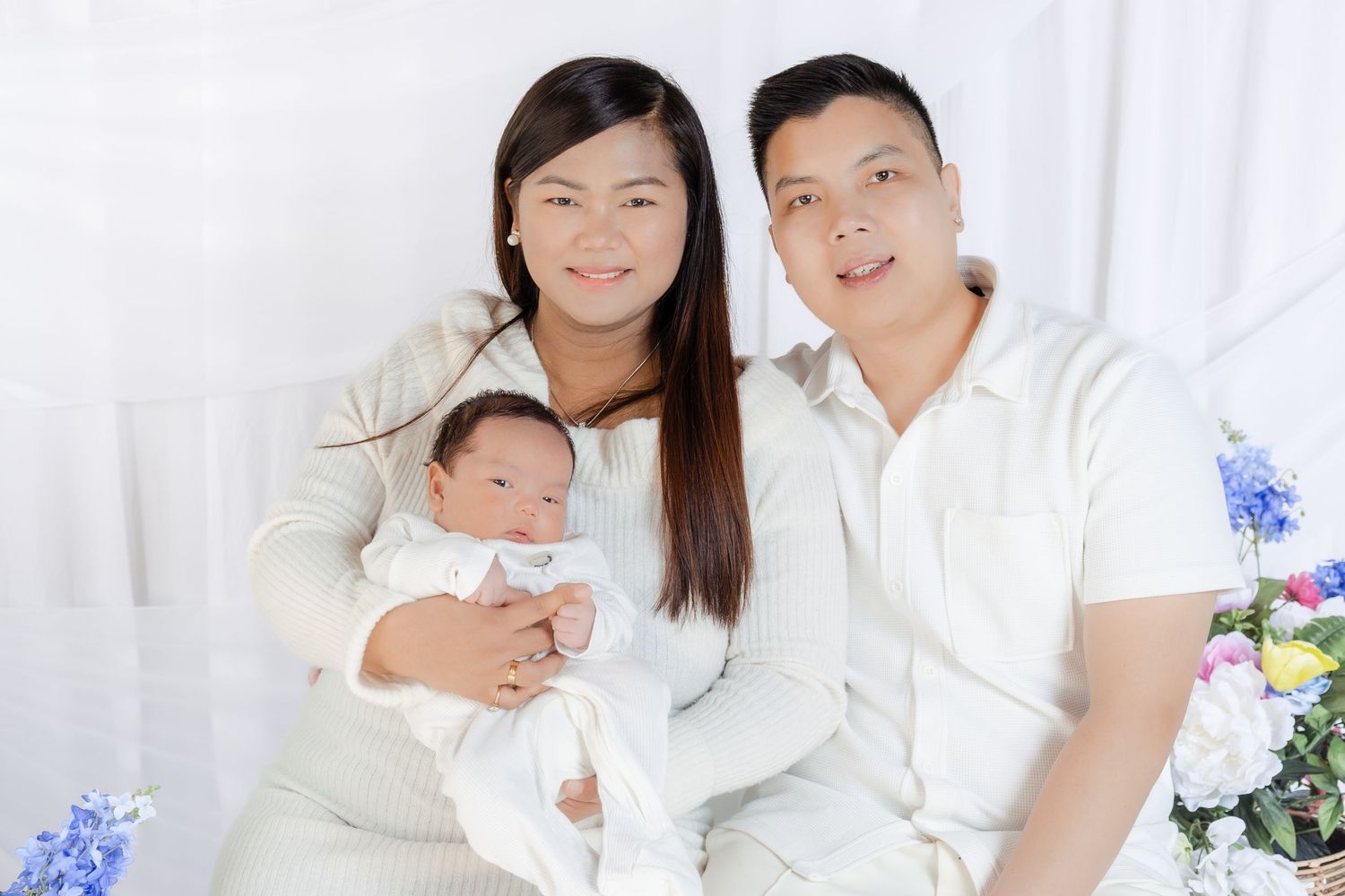 Happy Asian family portrait with mother holding newborn baby beside father, all dressed in white.