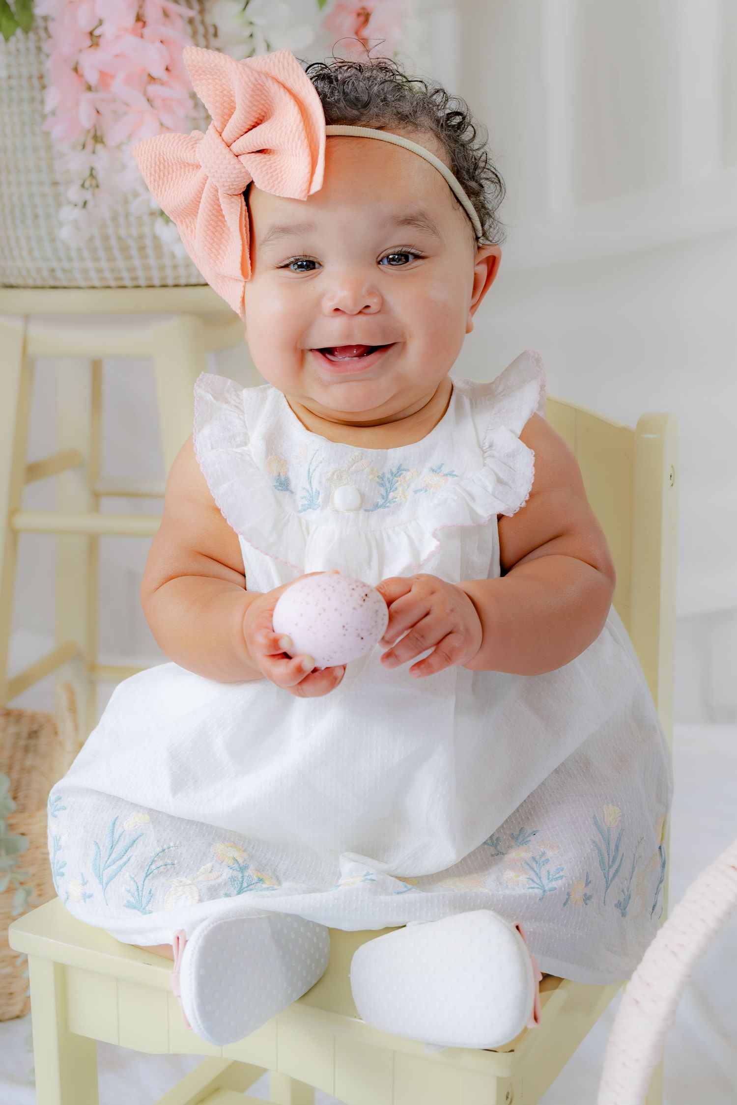 A joyful baby girl in a white embroidered dress and peach bow sits on a yellow chair holding a pink Easter egg.