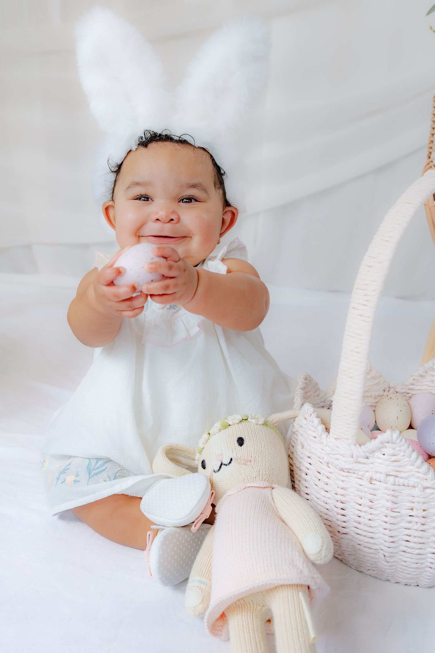 Happy baby girl in white dress with bunny ears holding Easter egg near basket.