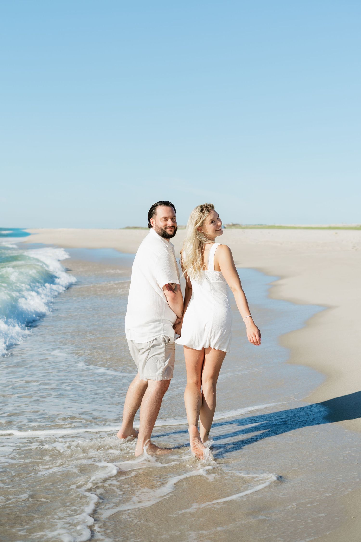 Couple in white outfits holding hands while walking barefoot along a sunny beach with gentle waves.