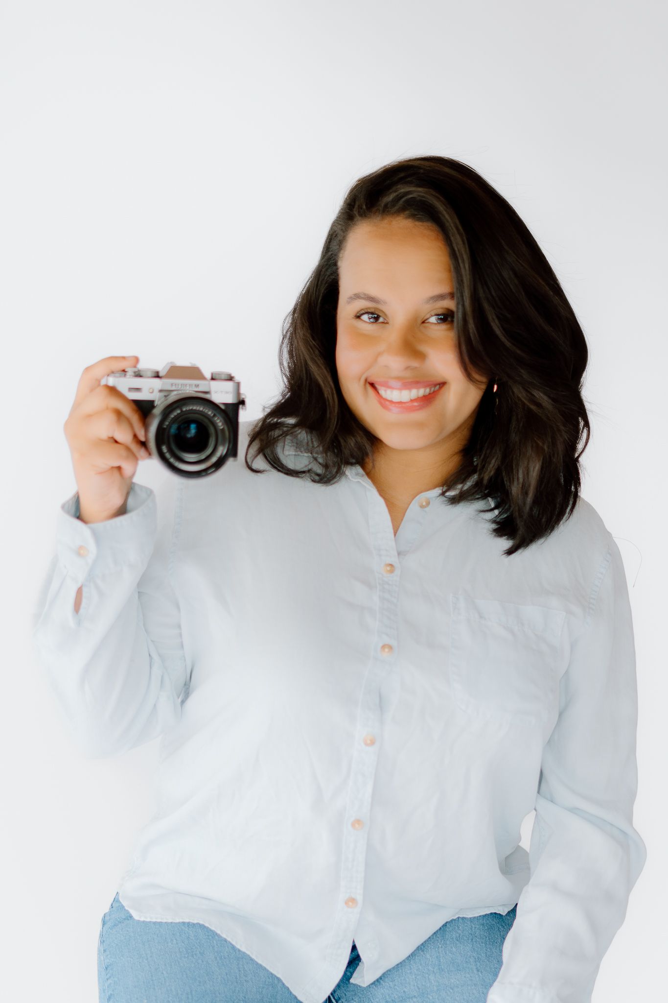 Smiling woman in white linen shirt holding a mirrorless camera against a white background.