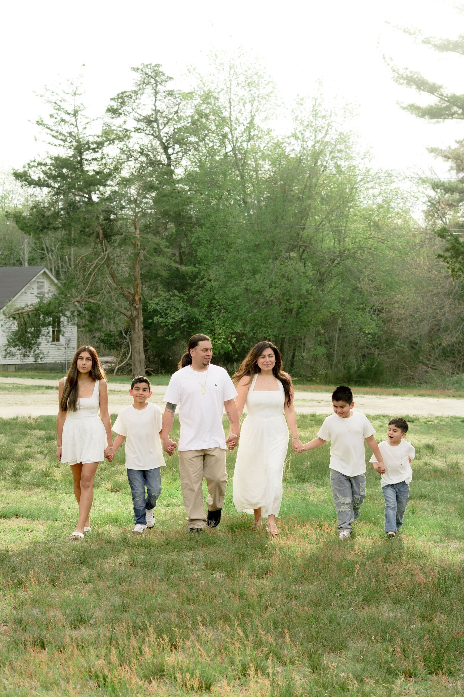 A family of five walks hand-in-hand through a green field, dressed in white and denim, surrounded by lush trees.