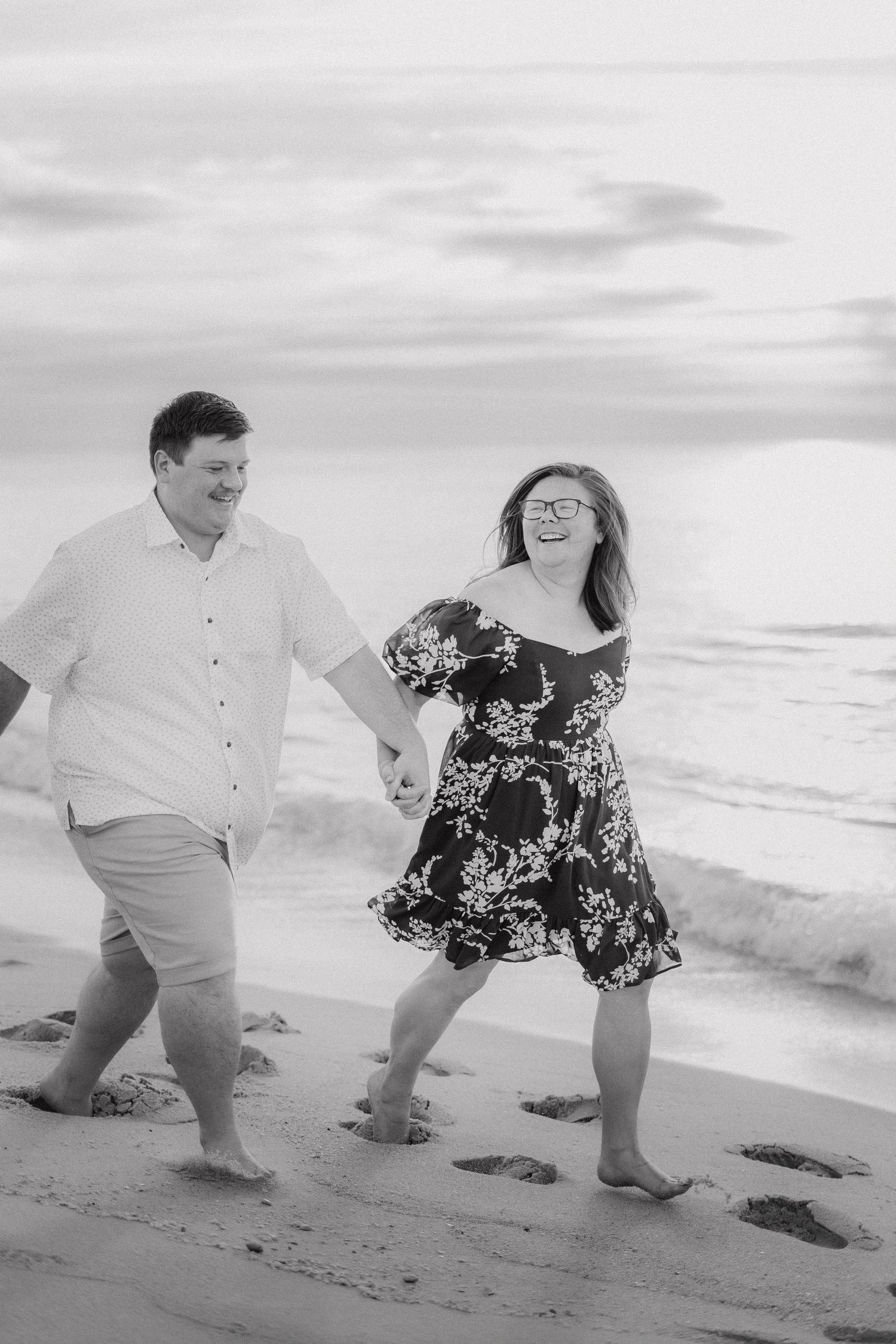 Black and white photo of a happy couple holding hands and laughing while walking barefoot on a sandy beach.