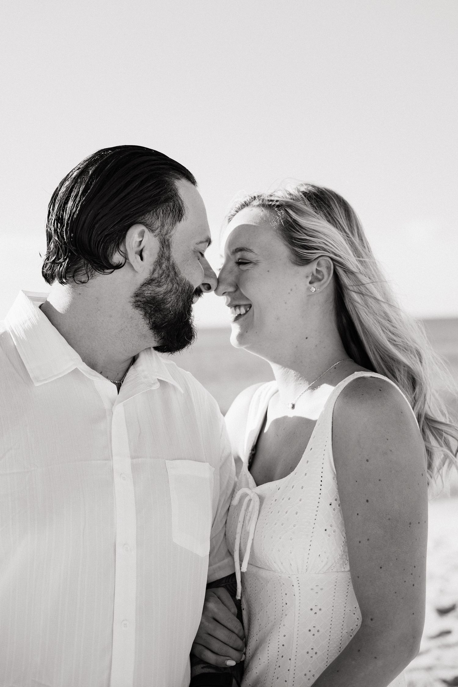 Black and white photo of a smiling couple almost kissing on a beach, woman in white dress, man in white shirt.