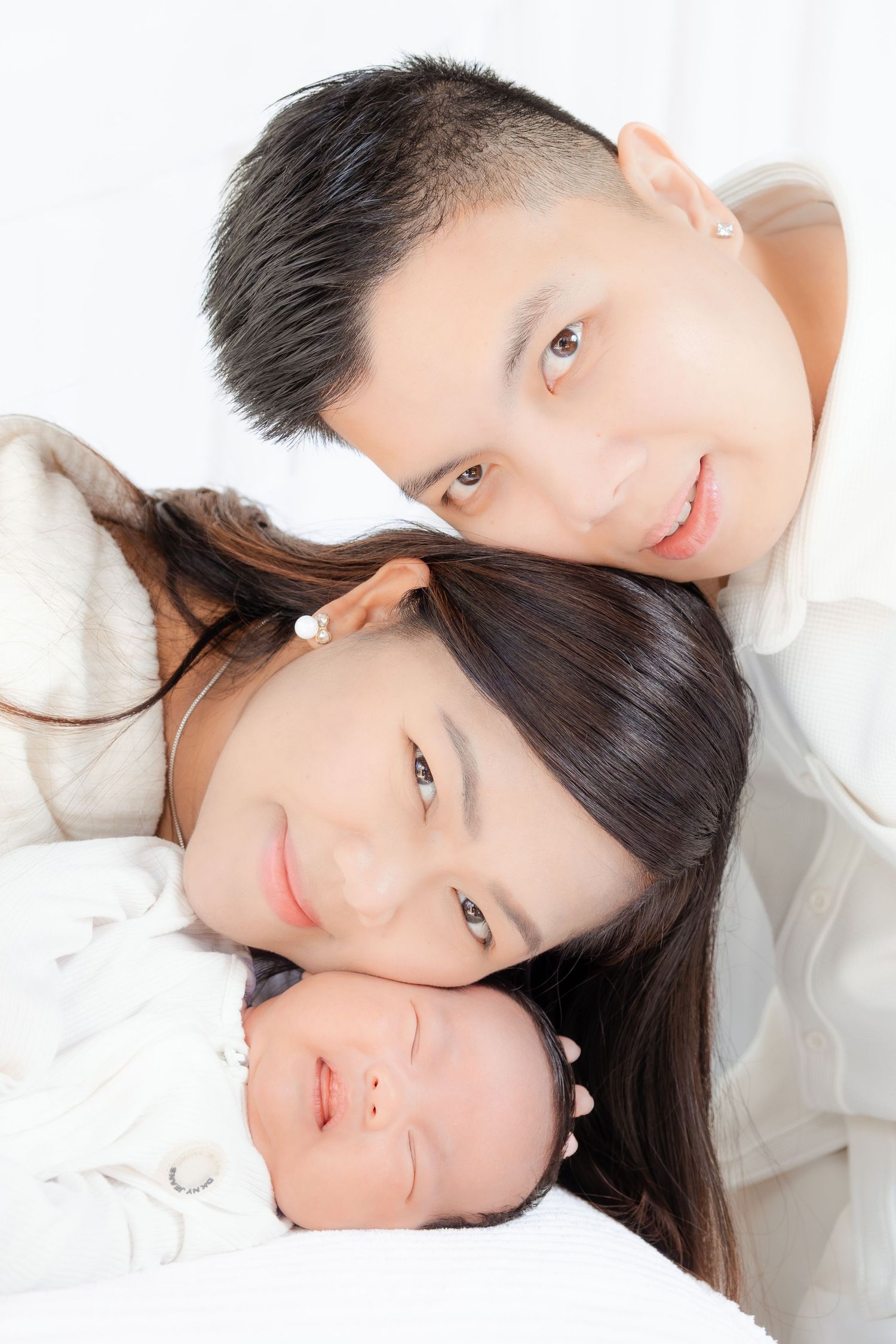 Asian family portrait with smiling parents lying close together with their sleeping newborn baby.