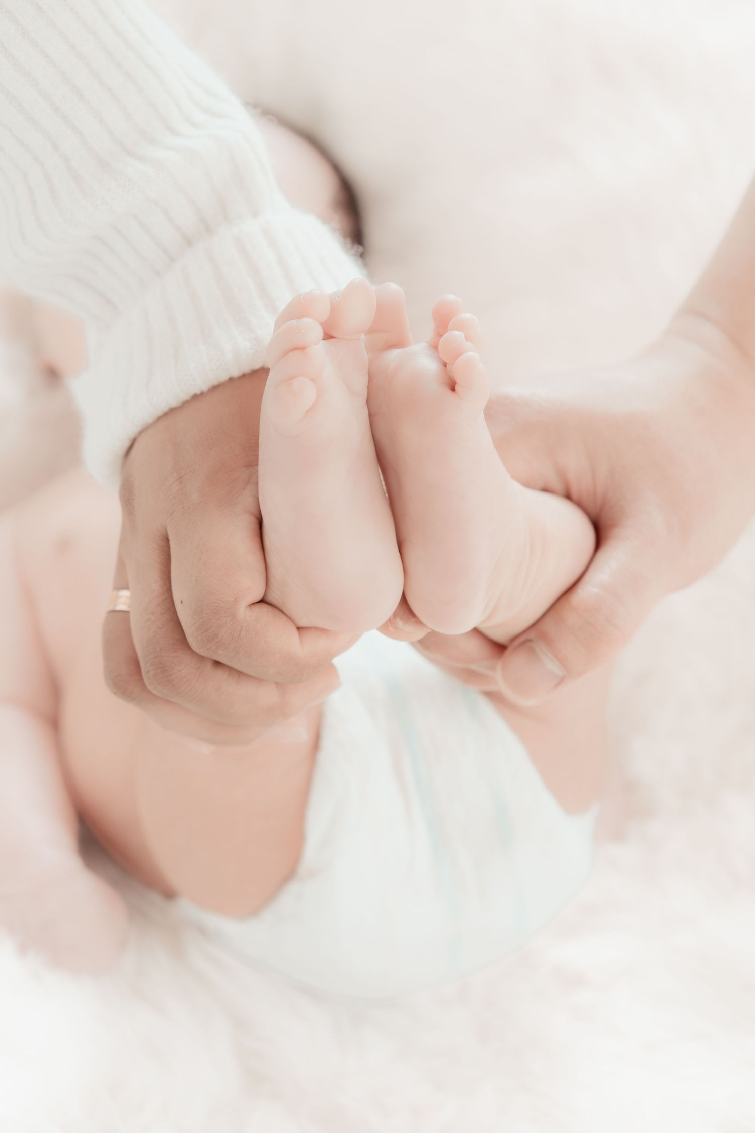 Adult hands gently cradling tiny newborn baby feet, wearing white knit outfit, soft white background.