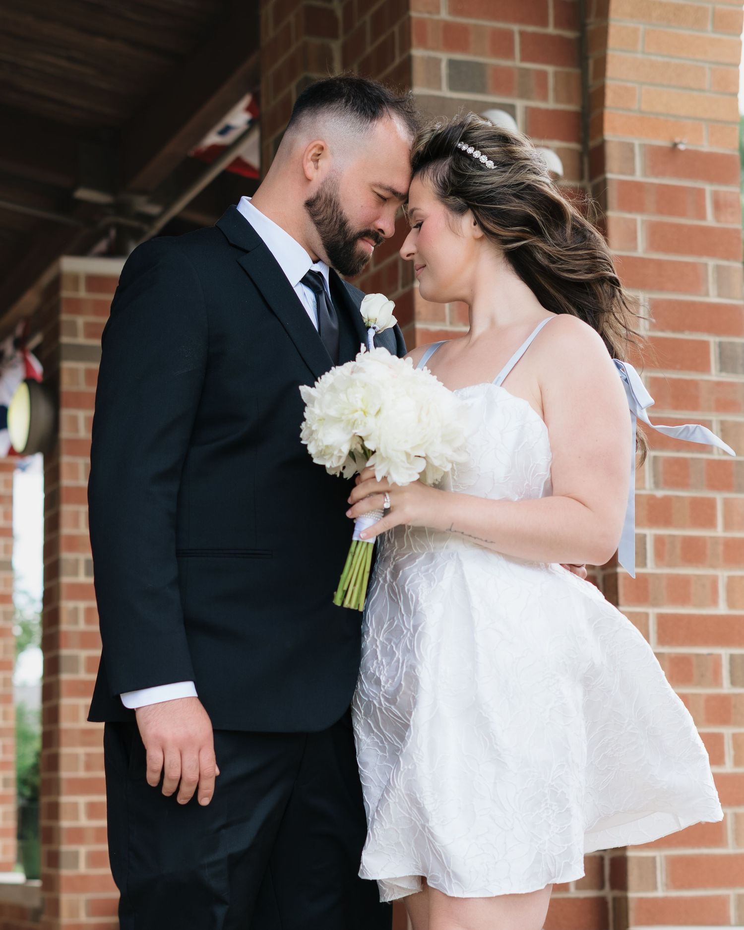 A couple in wedding attire sharing a romantic moment, bride holding white bouquet in short white dress, groom in black suit.