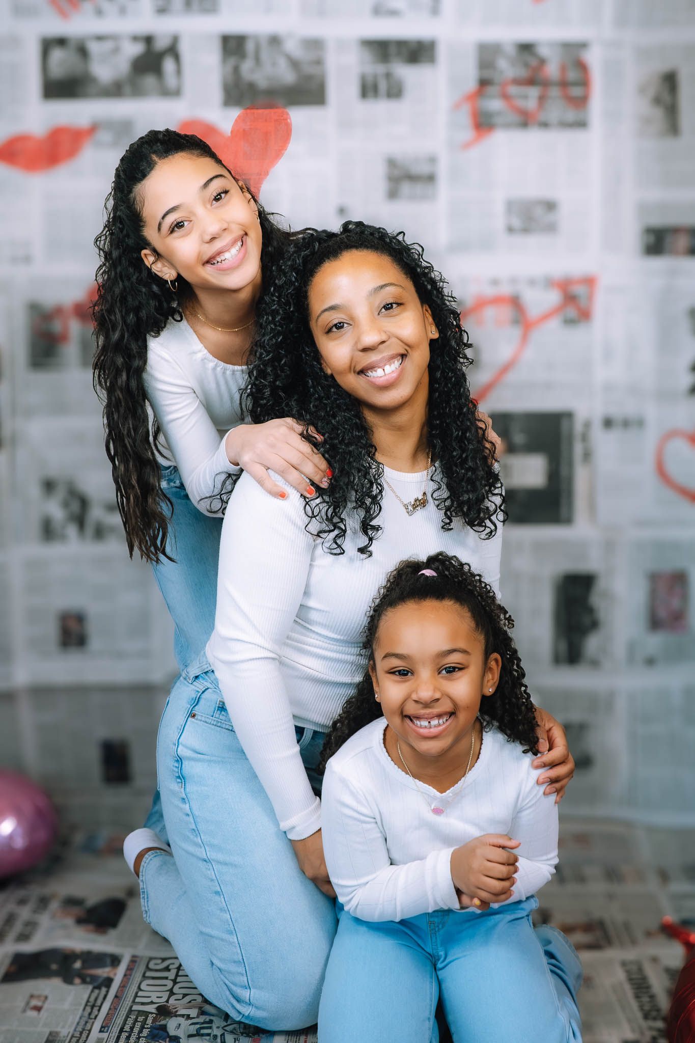 Three females with curly hair in matching white tops and jeans pose together in front of a newspaper backdrop.