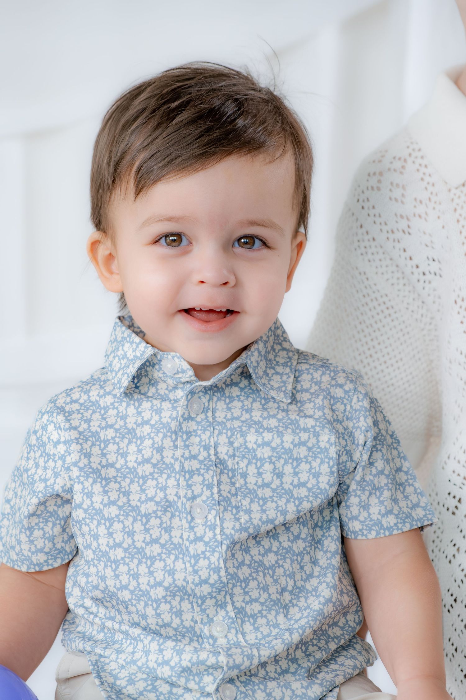 An adorable smiling toddler boy in a light blue floral button-up shirt poses against a white background.