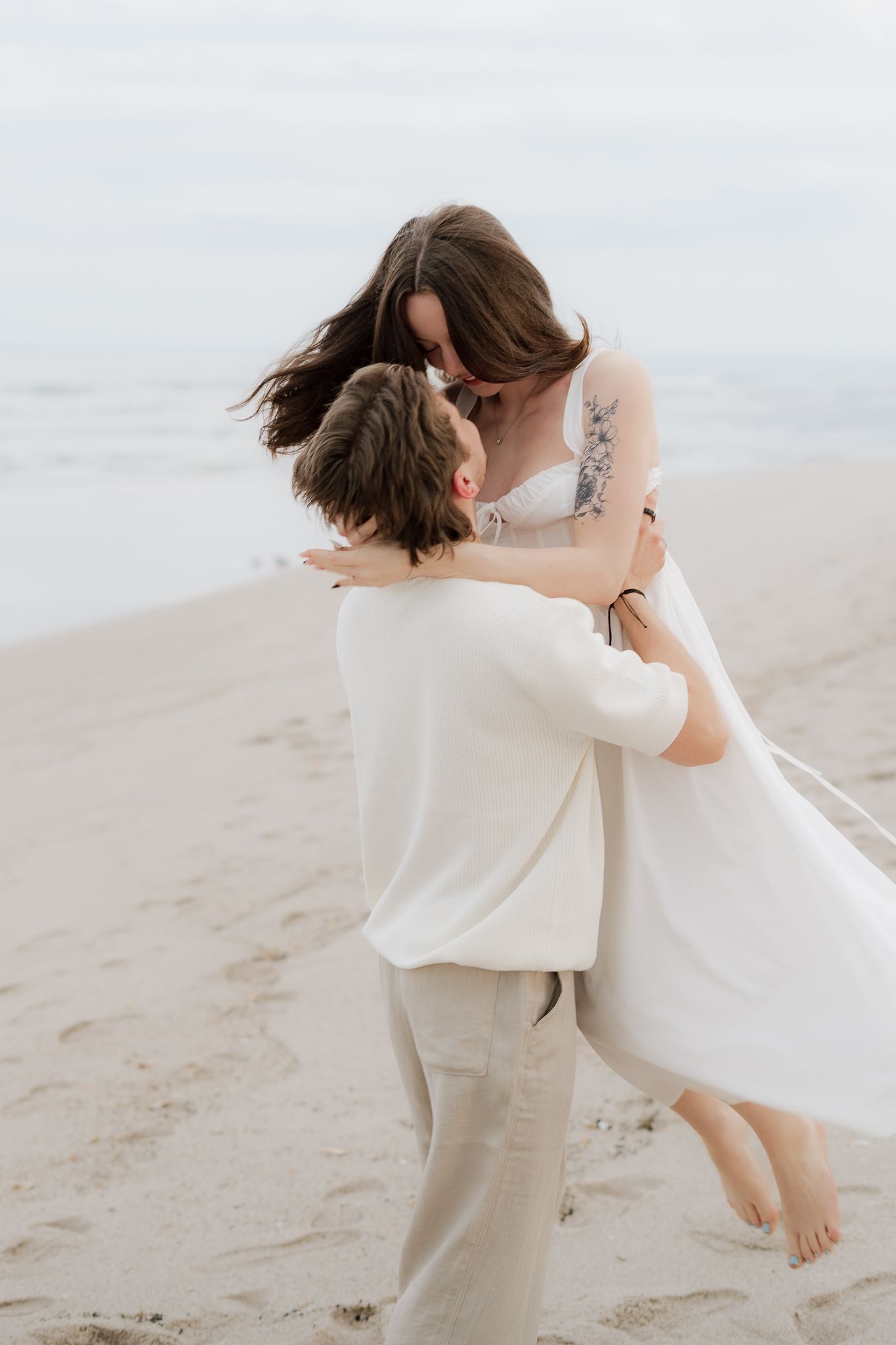 Groom lifting bride in white dress on a sandy beach during wedding photoshoot.
