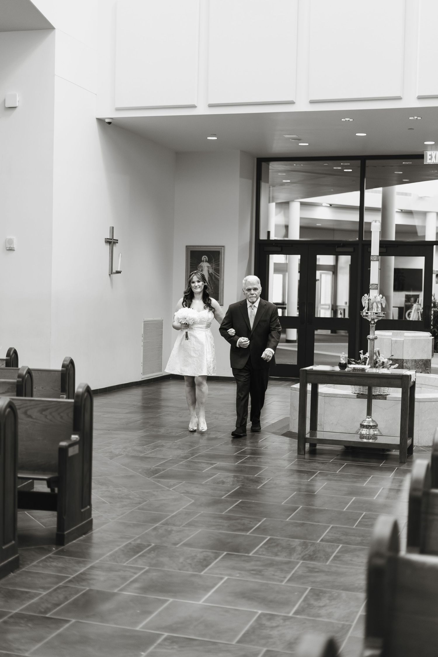 Black and white photo of bride in short white dress being escorted down church aisle by father on wedding day.