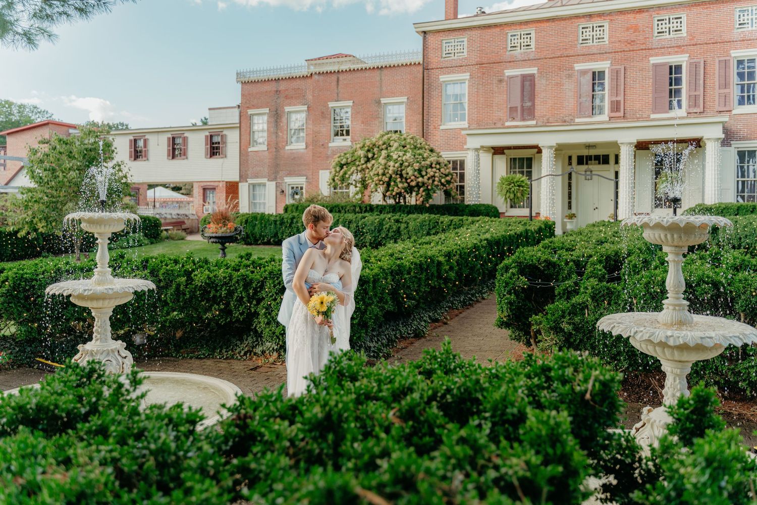 Bride and groom embrace in a formal garden with boxwood hedges and stone fountains outside a red brick estate.