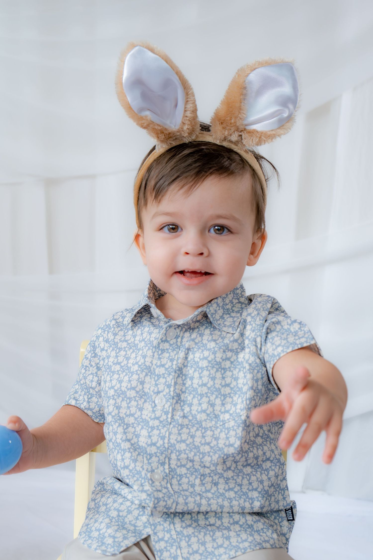 Toddler boy with bunny ears headband reaching toward camera in floral blue shirt.