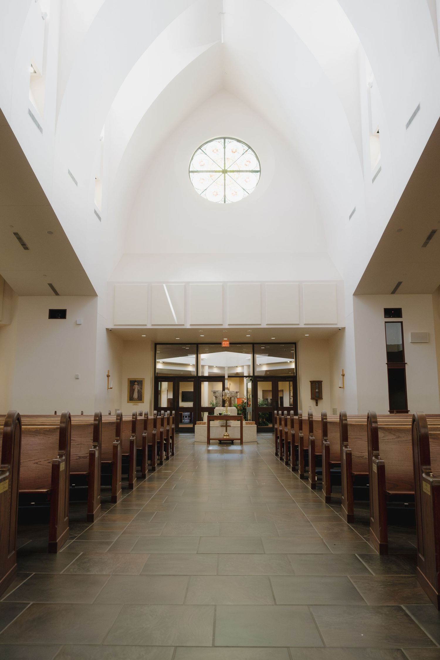 Bright modern church interior with white vaulted ceiling, wooden pews lining center aisle, and circular window above entrance doors.