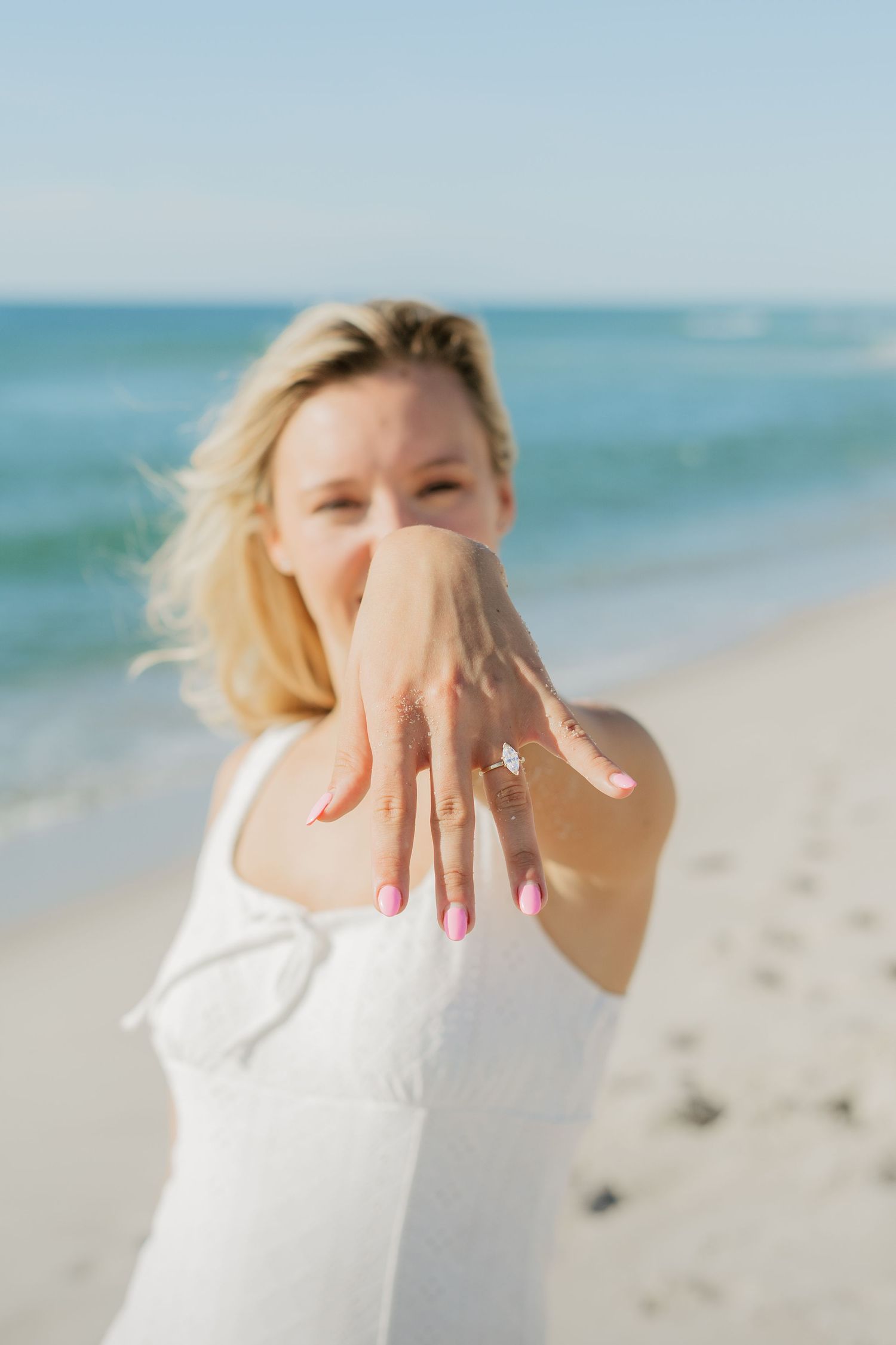 Smiling blonde woman in white dress showing off her diamond engagement ring on a sunny beach.