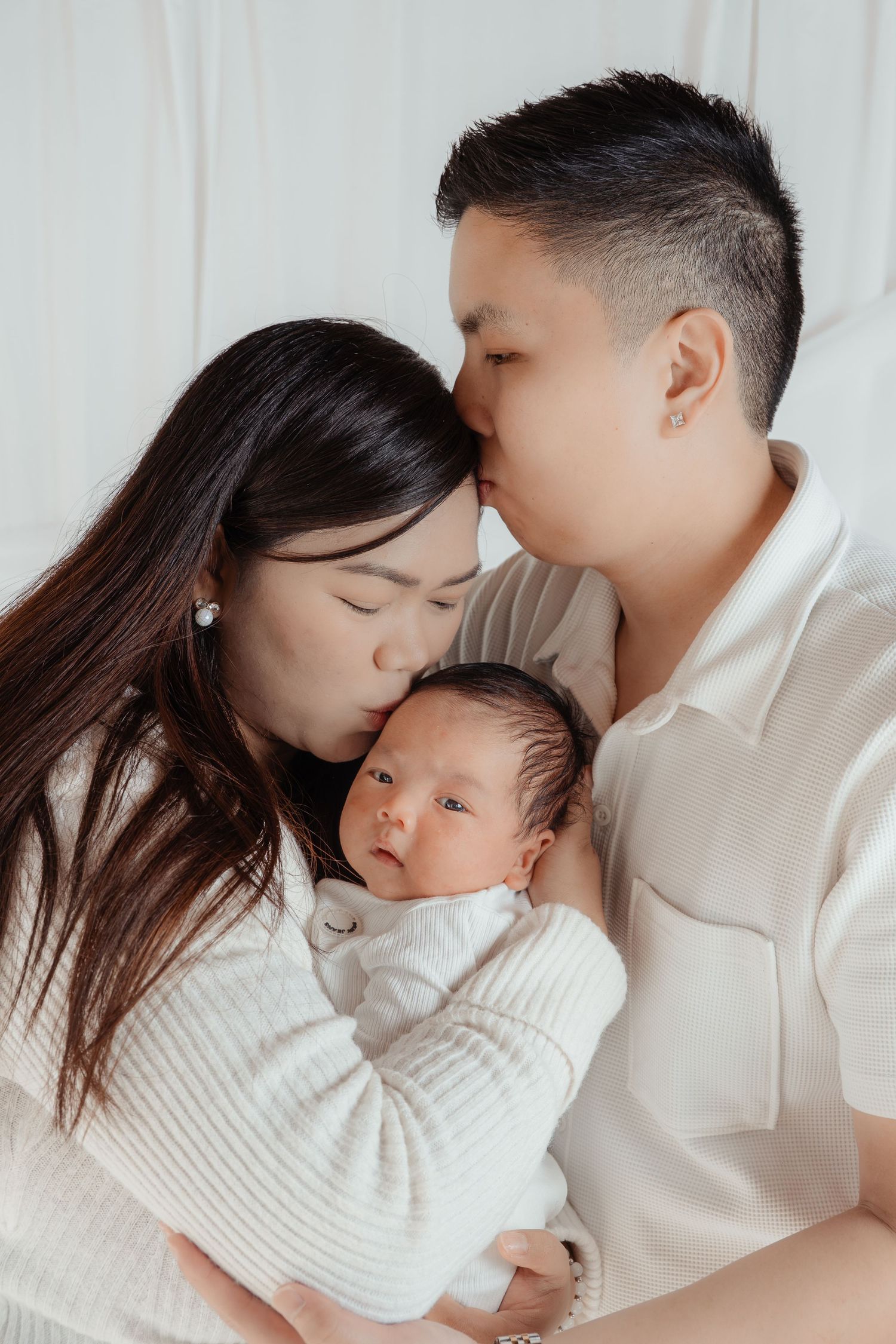 Two parents tenderly kissing their newborn baby wrapped in white, captured in a soft studio portrait.