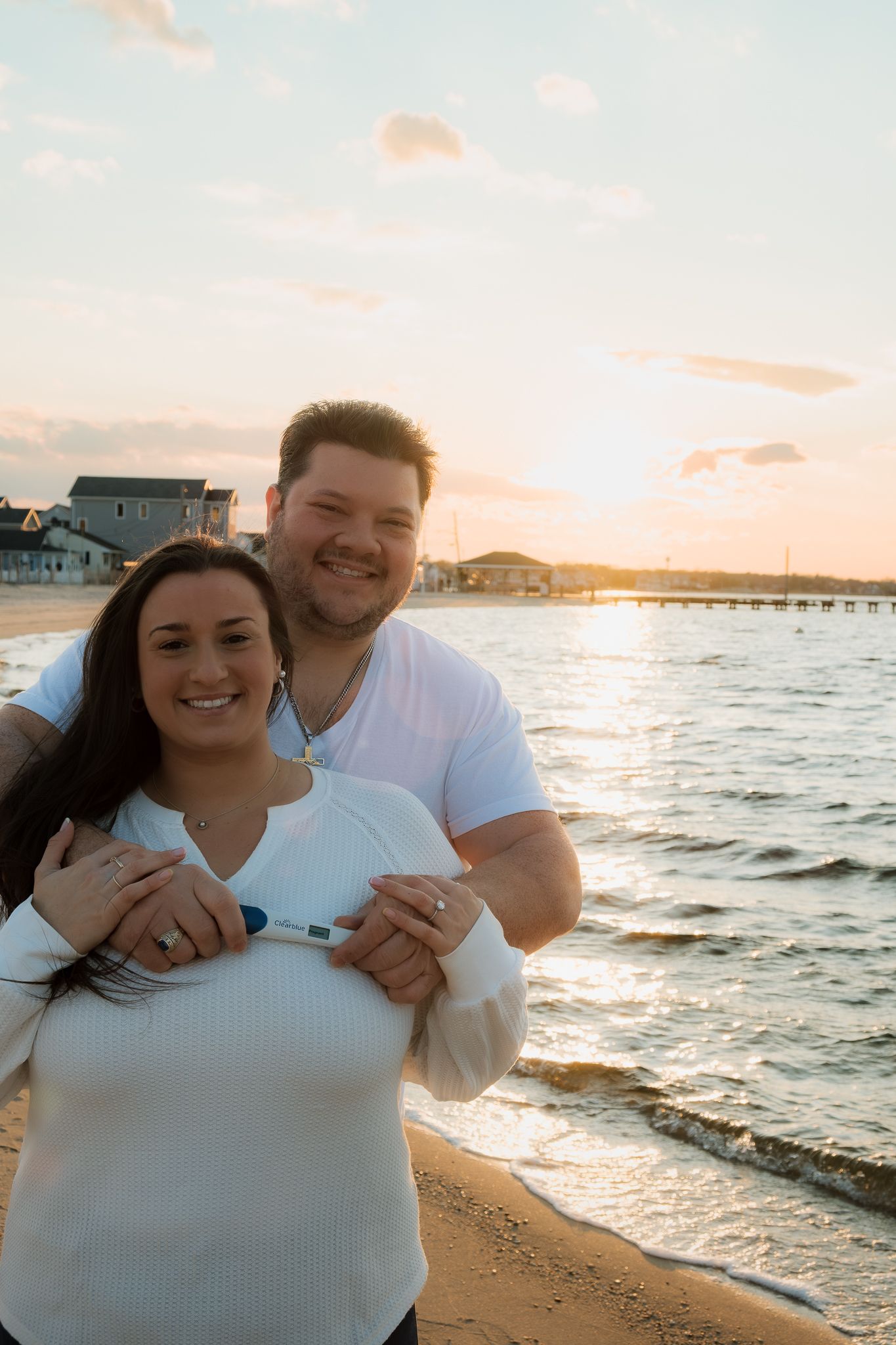 Happy couple embracing on a sandy beach at sunset, holding a pregnancy test, with a pier and waterfront homes in the background.