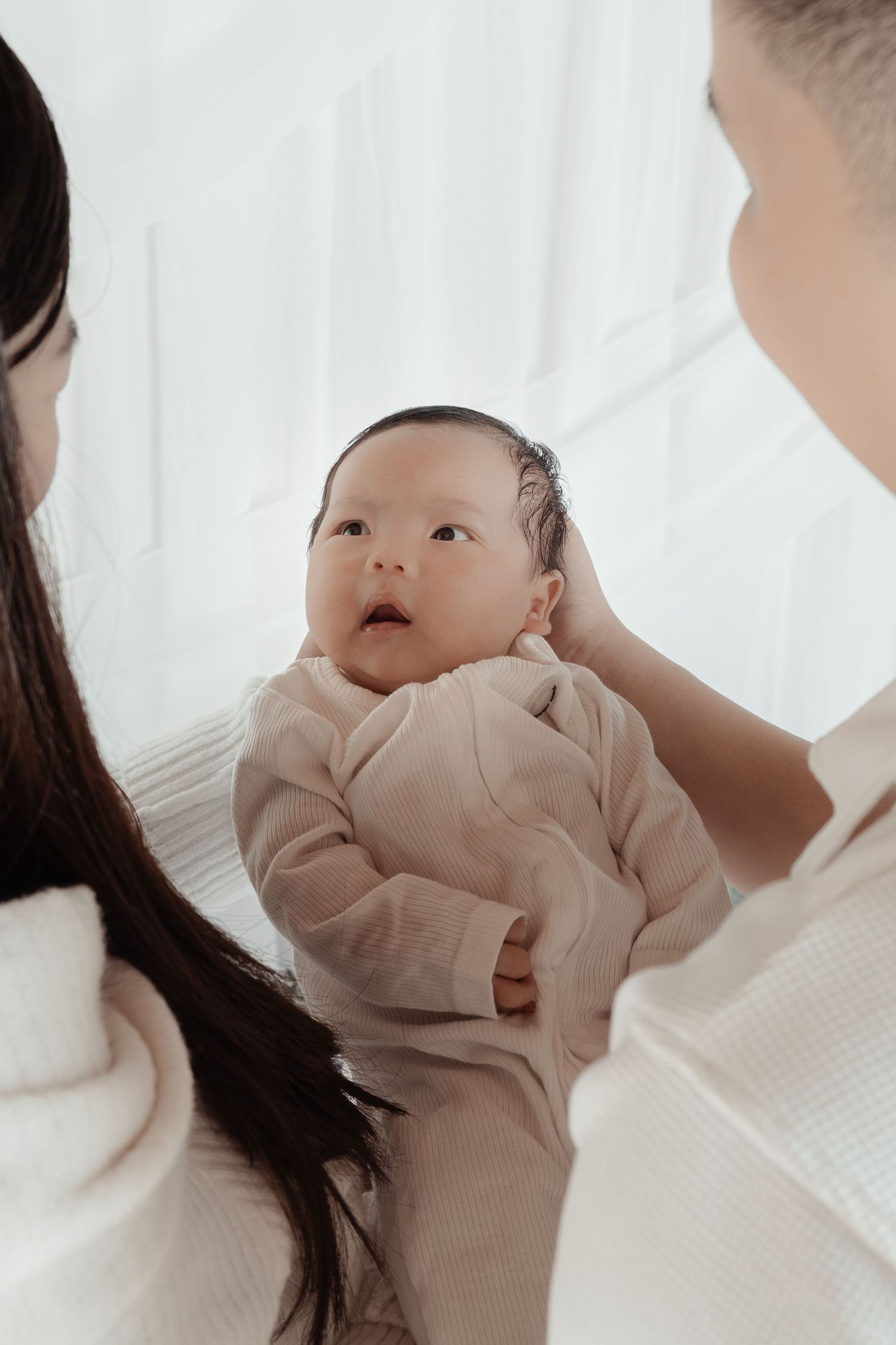 Parents holding their newborn Asian baby in a bright white room, baby wearing a beige onesie looking up curiously.