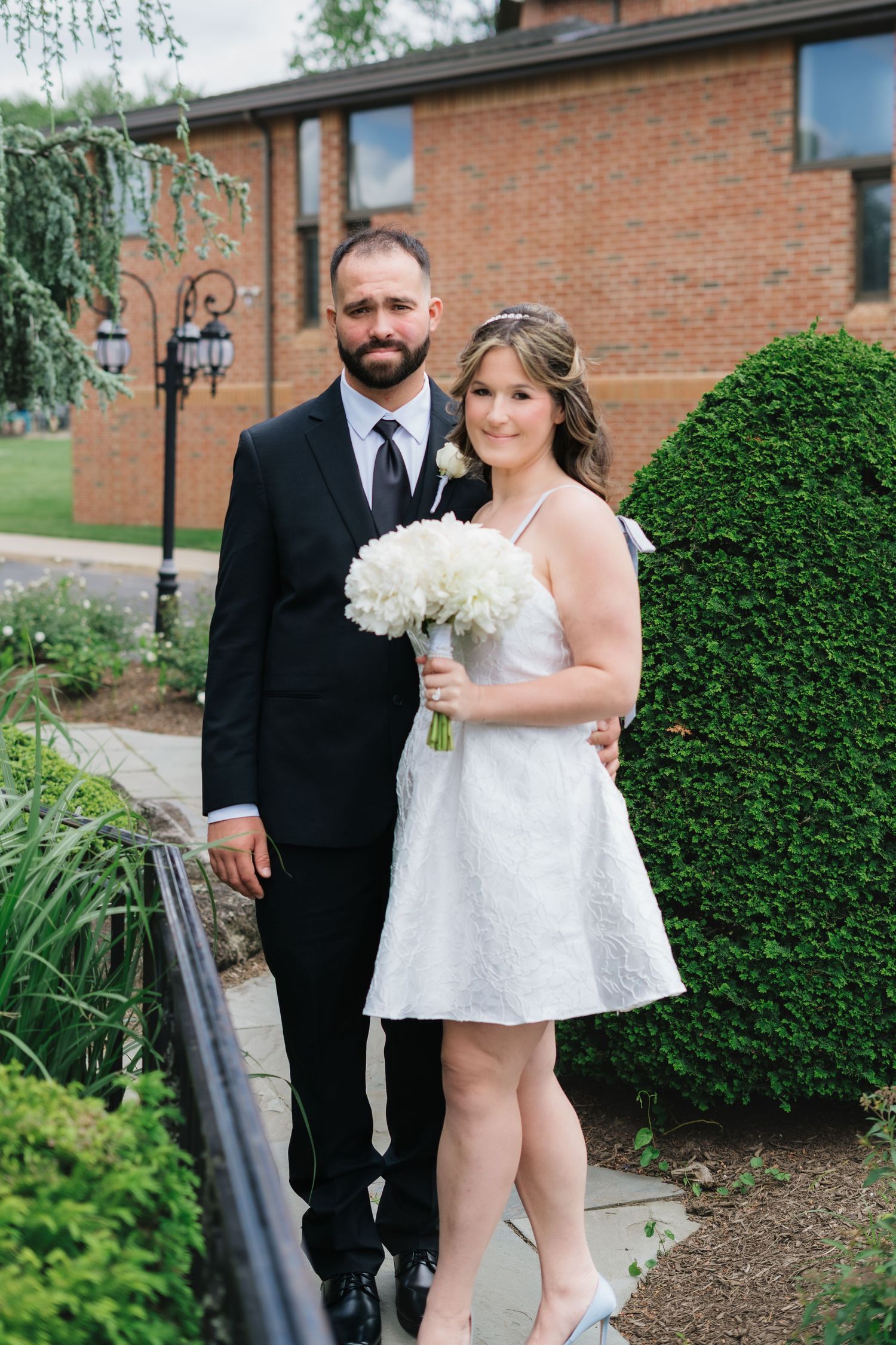Bride and groom pose together outdoors, bride in short white dress holding white peony bouquet.