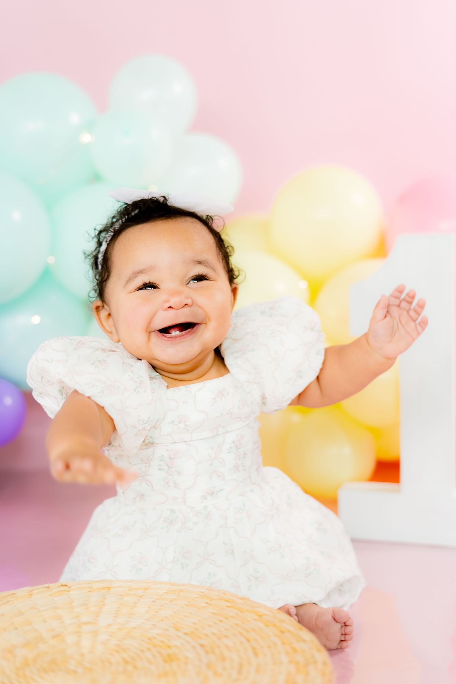 Joyful baby girl in white dress laughing with arms raised, surrounded by colorful pastel balloons at first birthday party.