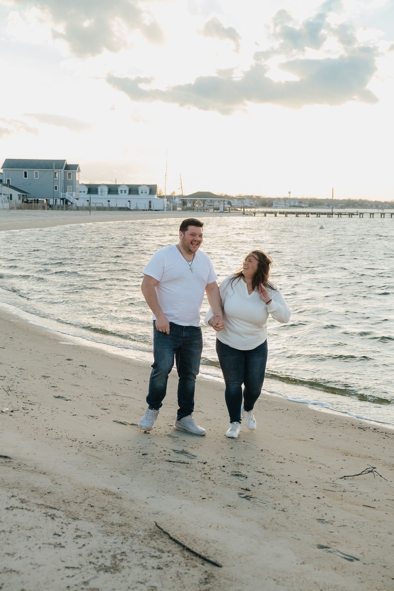 A happy couple in matching white tops and jeans walk hand-in-hand along a sandy beach at sunset.