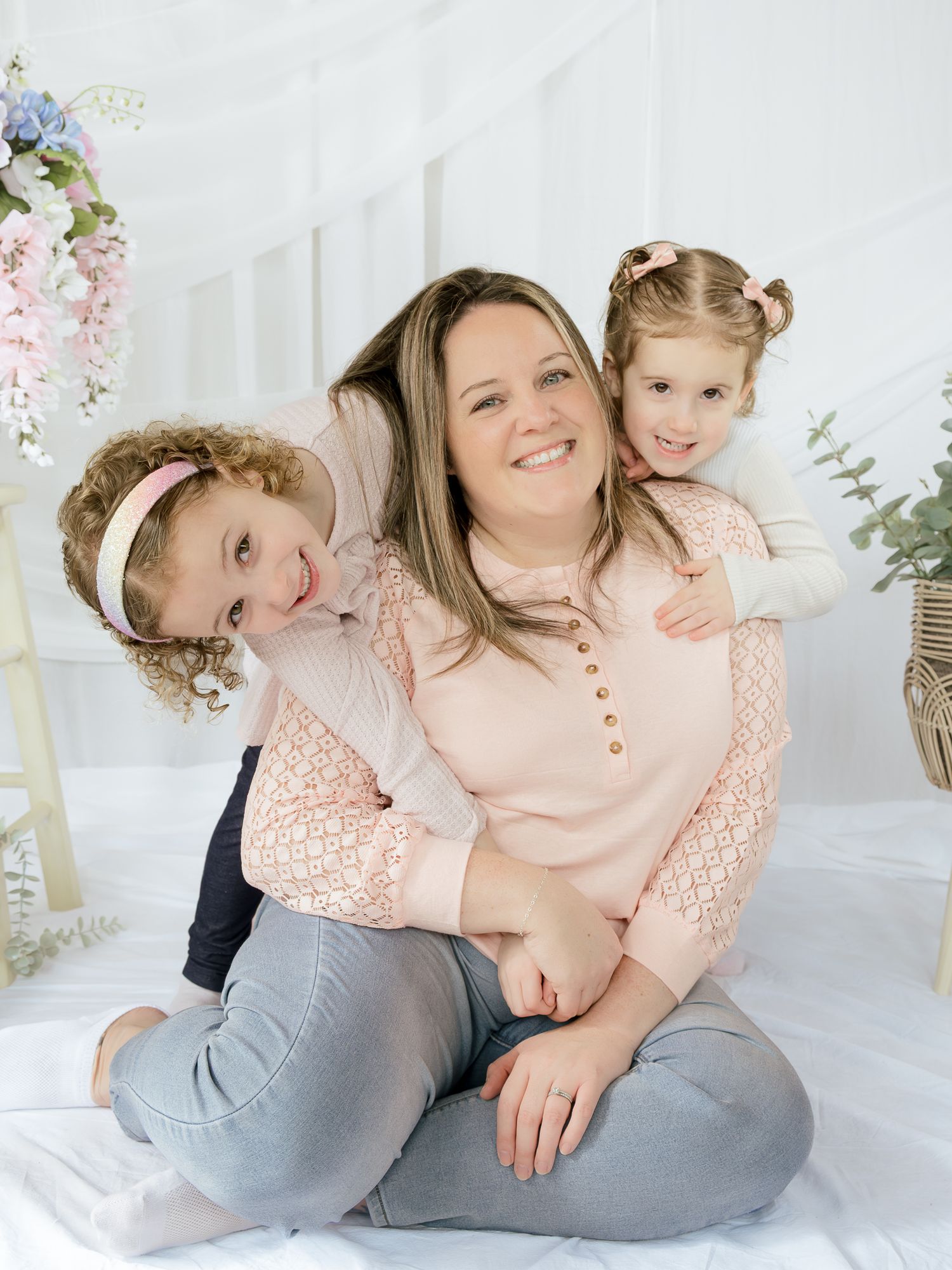 A mother sits on the floor hugging her two daughters in pink outfits in a bright white floral studio.