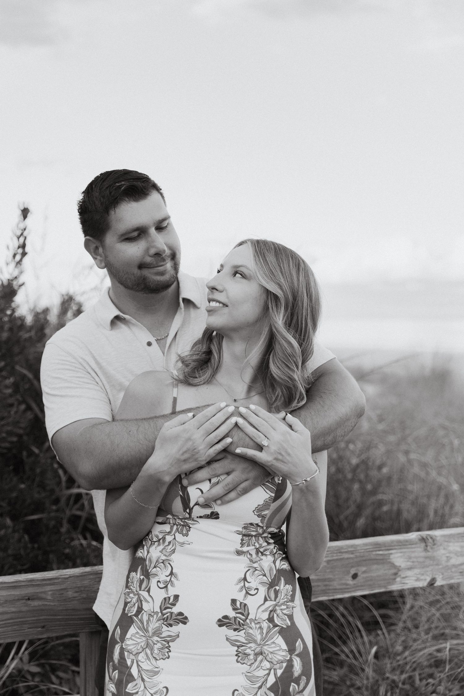 Black and white photo of a couple embracing outdoors, woman looking up smiling, man holding her from behind near a wooden fence.