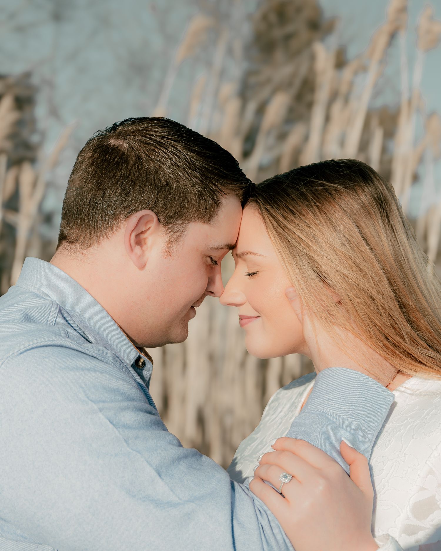 Romantic couple touching foreheads outdoors, woman wearing engagement ring, soft natural background with tall reeds.