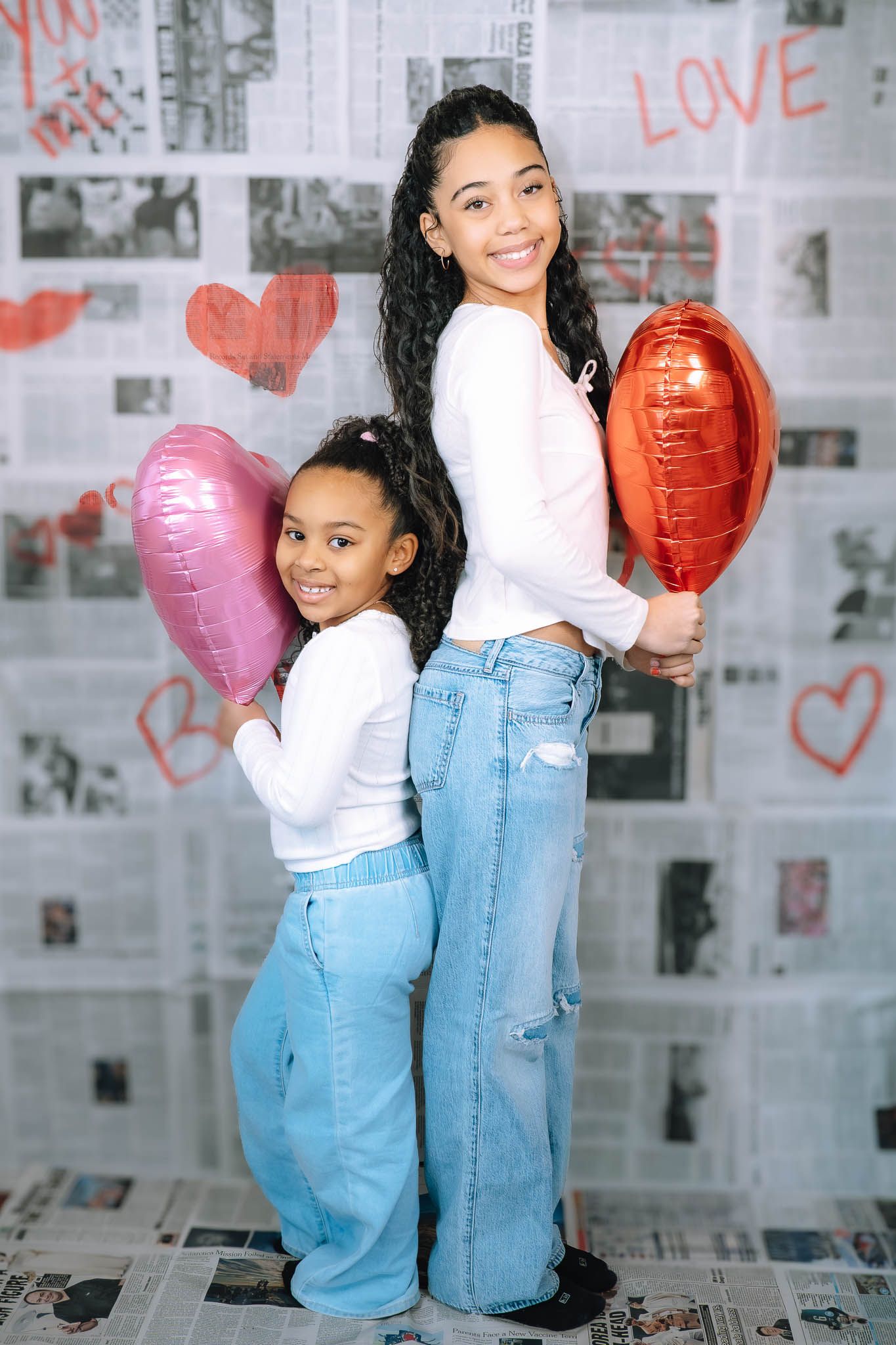 Two sisters posing back-to-back holding pink and red heart balloons in front of Valentine's Day newspaper backdrop.