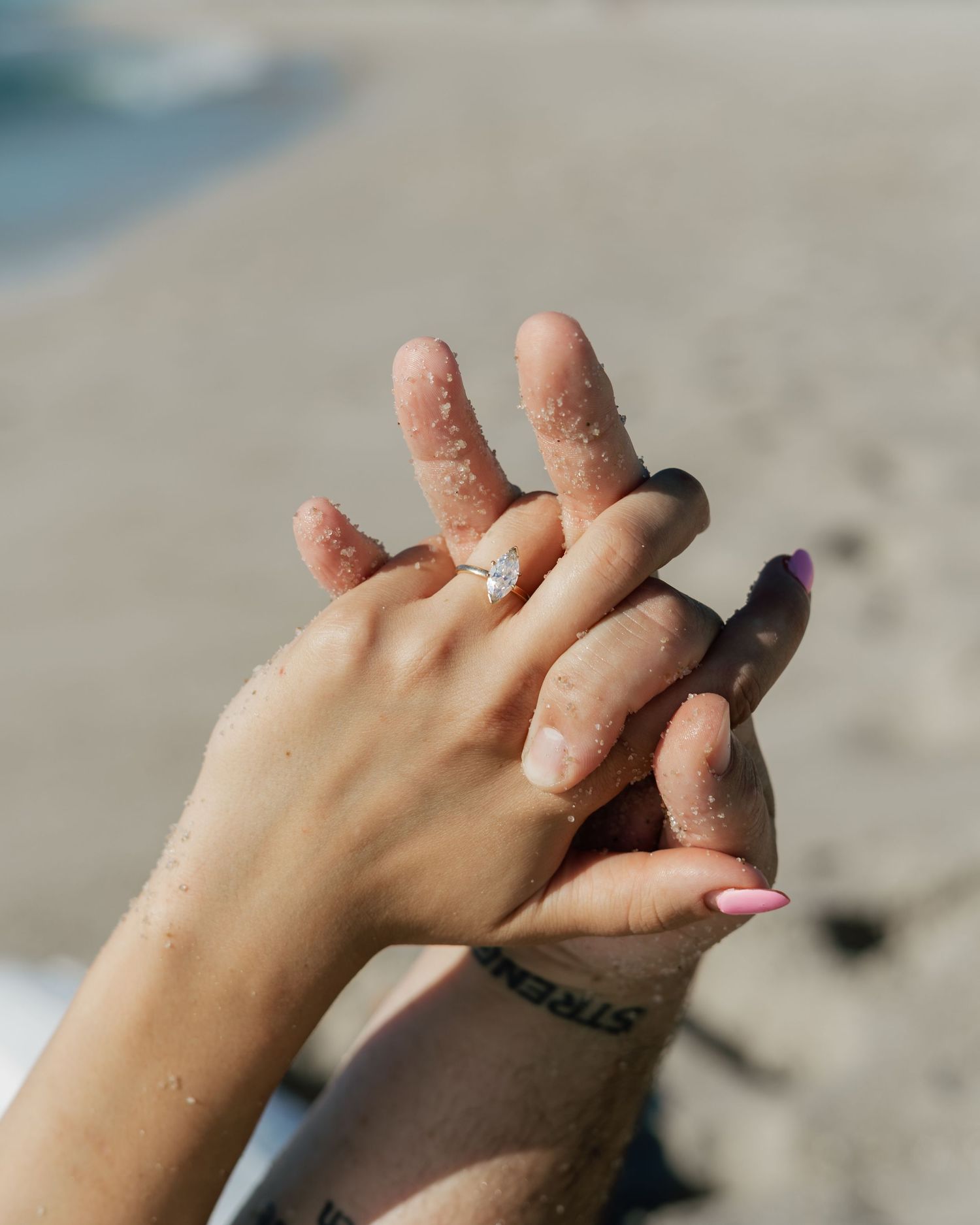 Two sandy hands touching on a beach, one wearing a diamond engagement ring with pink and purple nail polish.