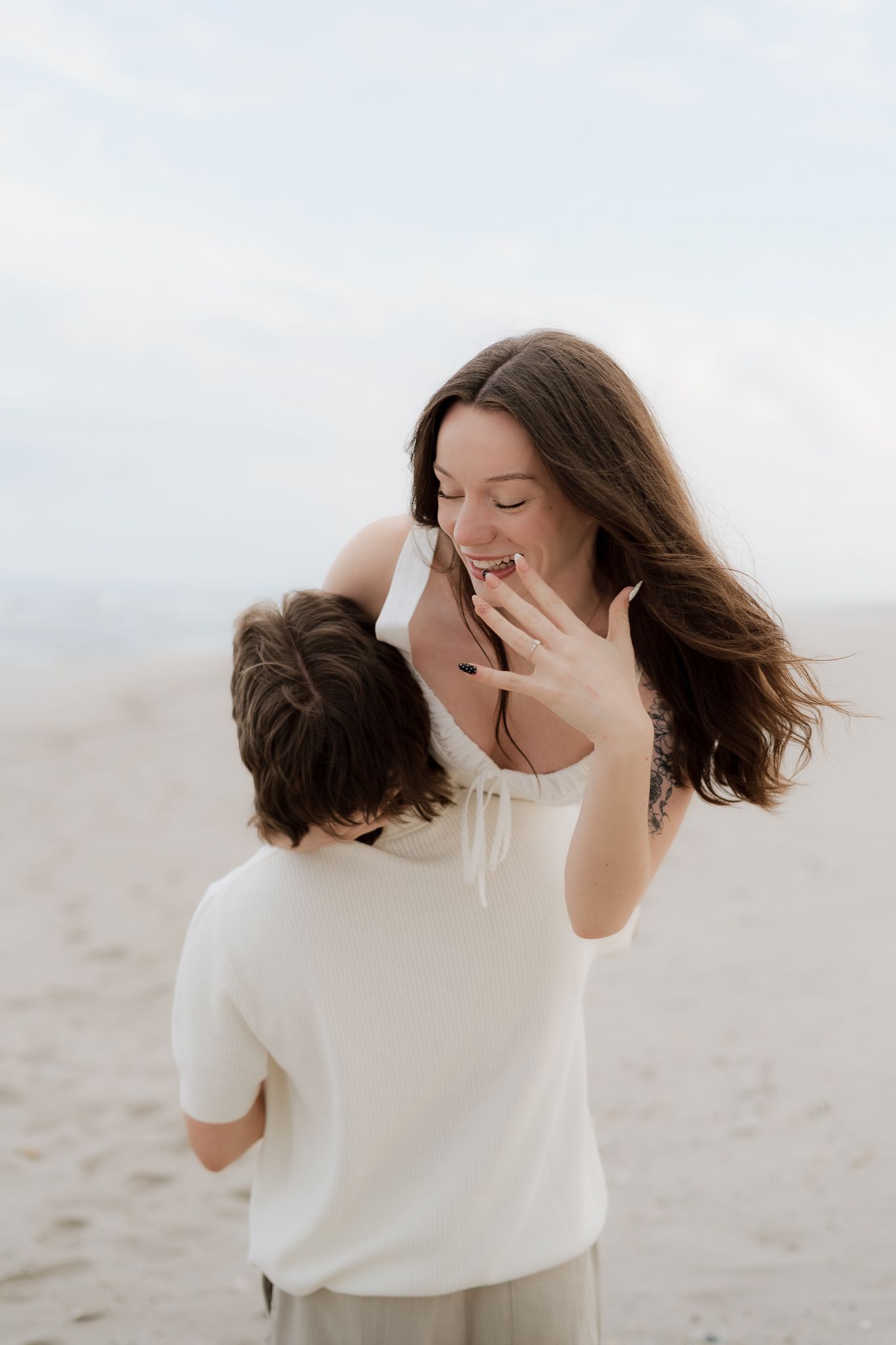 Joyful woman shows engagement ring on sandy beach, covering mouth in surprise while embraced by partner in white.