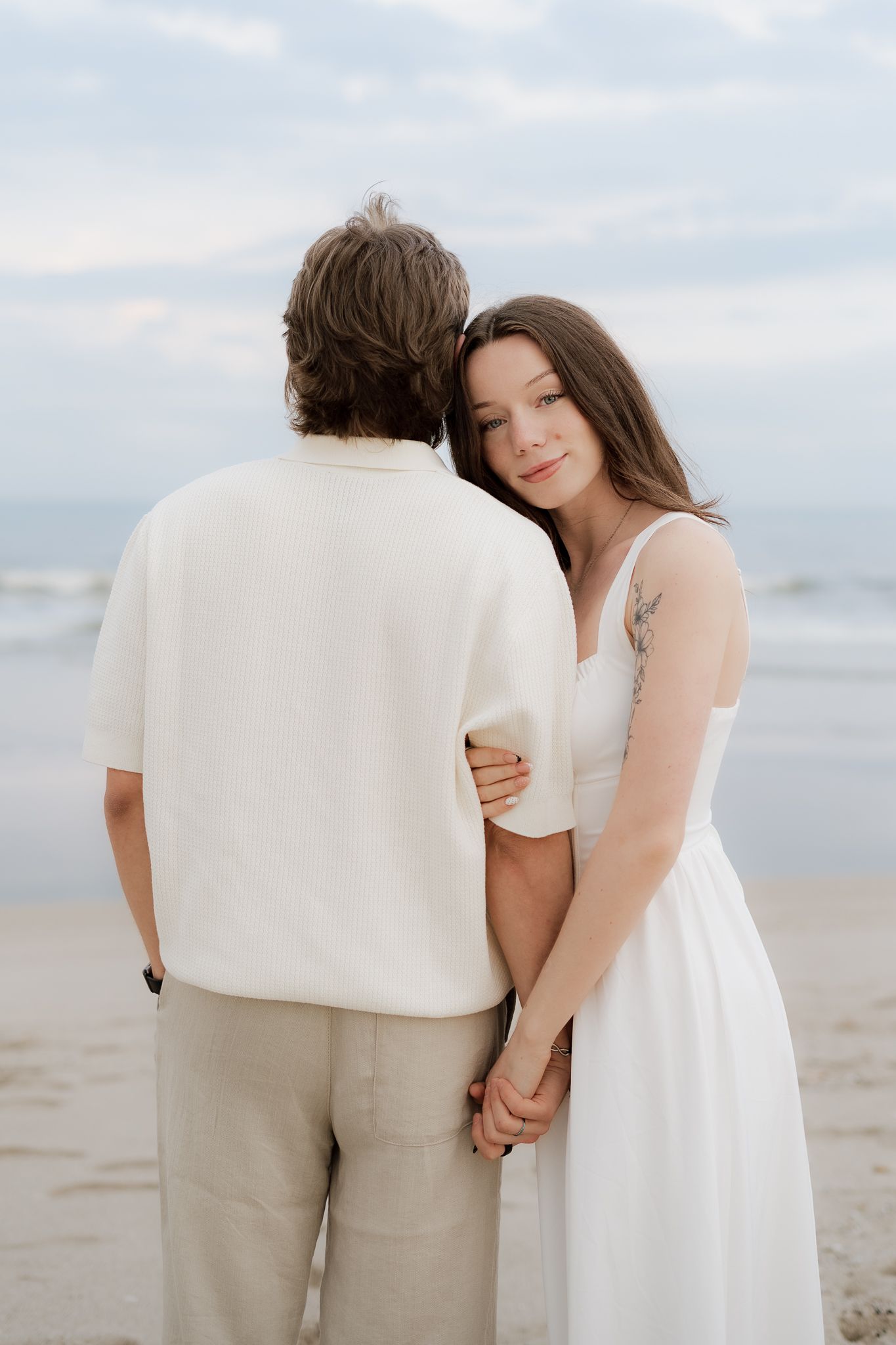 A couple in white outfits embracing on a beach, woman with tattoo resting head on man's shoulder.