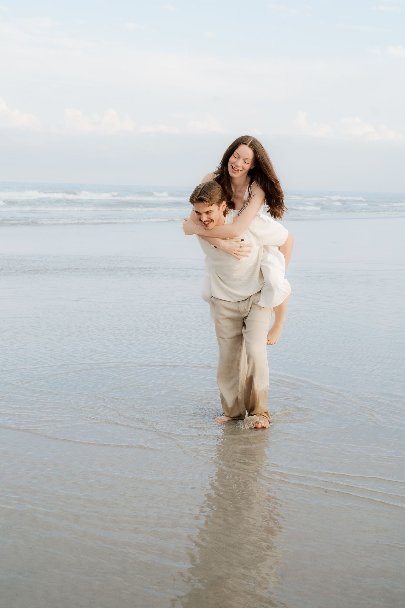 A couple laughing on the beach, man giving woman a piggyback ride in shallow ocean water.