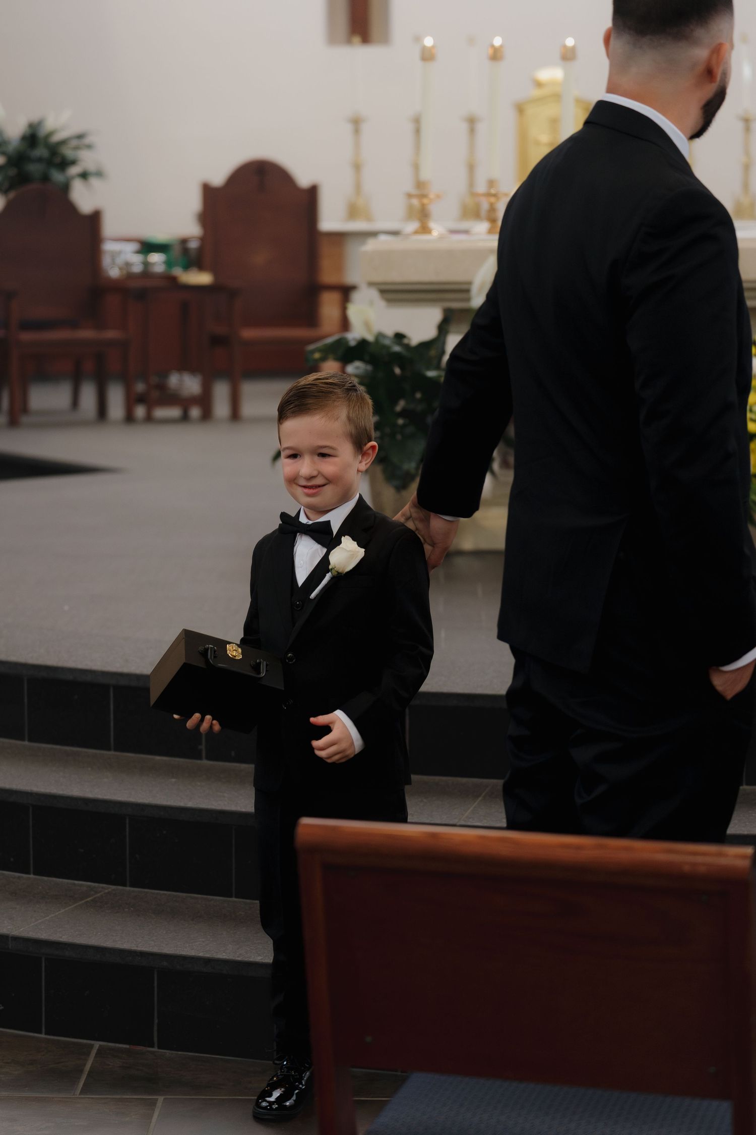 Adorable young ring bearer in black tuxedo with white boutonniere smiles while holding a book during a church wedding ceremony.