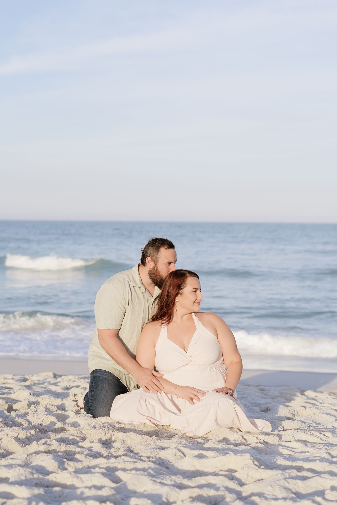 A couple sits together on a sandy beach, the man embracing the pregnant woman as gentle ocean waves roll in behind them.