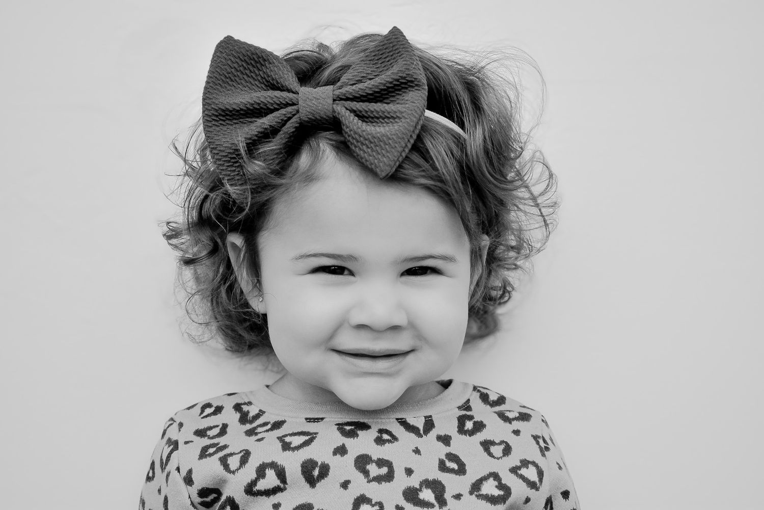 Adorable curly-haired toddler girl smiling in black and white portrait wearing a bow headband.