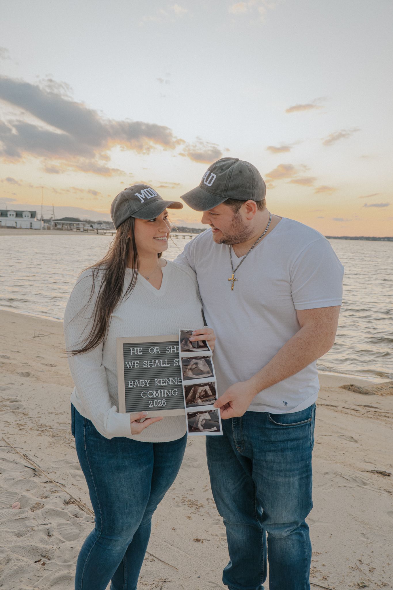 Couple holding pregnancy announcement sign on beach at sunset, revealing Baby Kenne coming 2025.