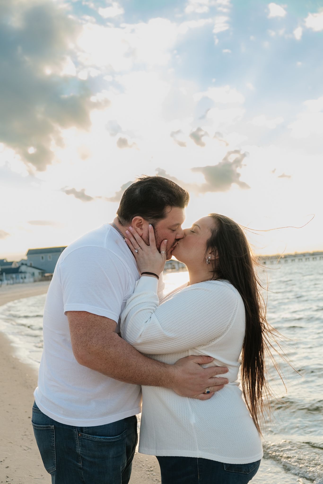 Couple sharing a romantic kiss on the beach at sunset, wearing white tops and jeans.