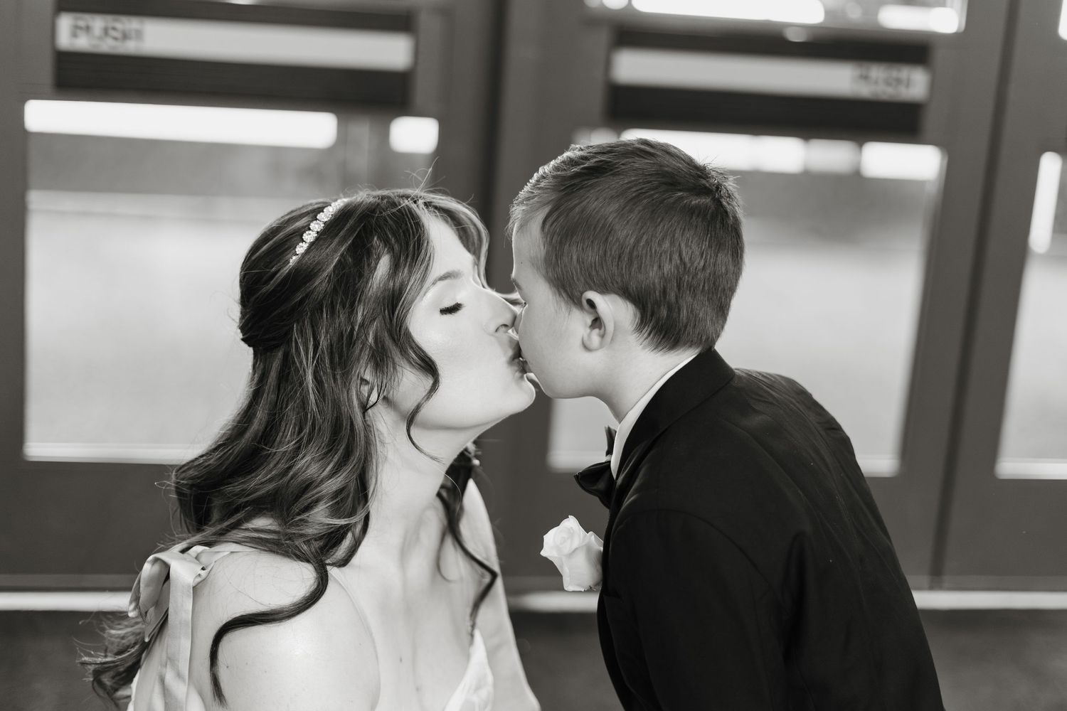 Black and white photo of bride kissing young boy in black tuxedo near push door at wedding venue.