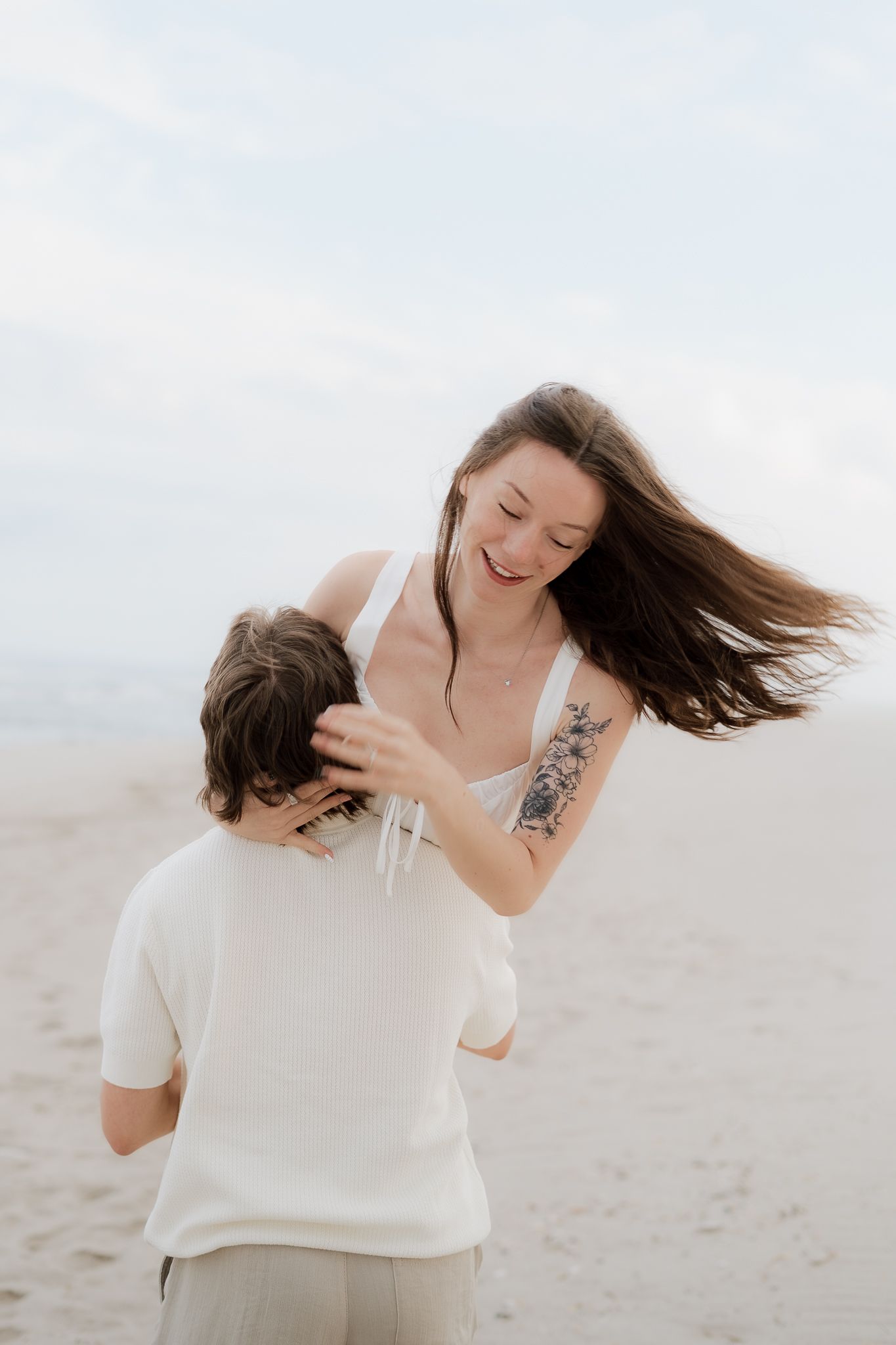 Laughing woman with flowing hair and floral tattoo playfully touches man's head on a sandy beach in matching white outfits.